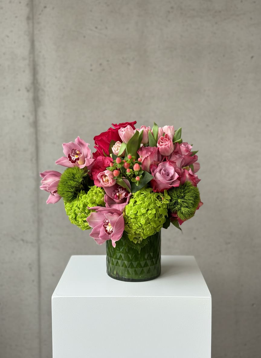 Eternal Elegance - “Eternal Elegance” is refined and striking, designed with a polished, modern touch. Lush green hydrangeas and green trick dianthus (green ball) form a rich, textured base, giving the arrangement depth and structure. Lavender and hot pink roses add vibrant contrast, blending soft romance with bold color.  Light pink tulips bring graceful movement, their smooth petals curving gently among the blooms, while mauve cymbidium orchids introduce an exotic, sophisticated flair. Pops of pink hypericum berries add subtle detail and a hint of shine.  Arranged tightly in a wide, diamond-embossed cylinder vase, the stems are hidden behind a sleek green leaf lining, creating a clean, seamless look. The overall design feels luxurious, balanced, and timeless—true to its name, “Eternal Elegance.”