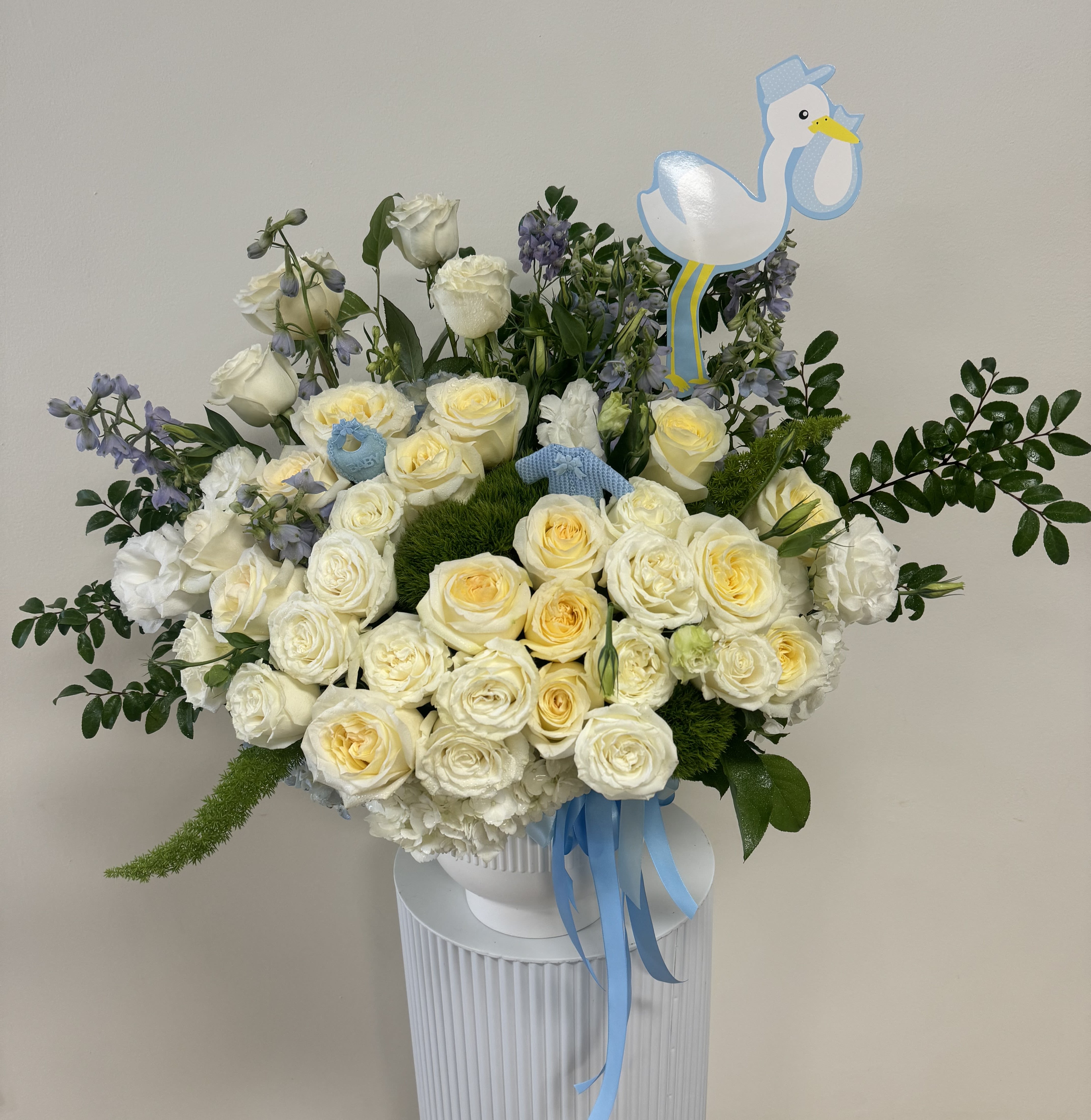 White and pale yellow roses in a vase with blue ribbon and a stork topper
