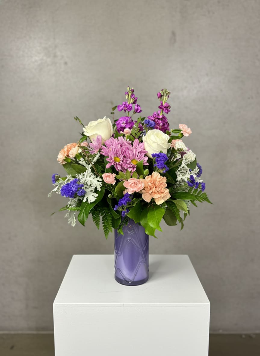 Sweet Serenity - “Sweet Serenity” is a soothing blend of soft color and gentle texture, arranged in a dark lavender vase with an elegant swirl design that adds depth and charm. Purple stock rises through the arrangement, bringing height and a light, calming fragrance.  White roses offer a classic, graceful touch, while pink carnations and mini carnations create a full, ruffled softness throughout. Lavender daisies add a cheerful, delicate feel, and accents of purple statice bring subtle pops of deeper color and texture.  Dusty miller weaves through the blooms with its silvery foliage, giving the arrangement a soft, velvety finish. Designed full and all around, “Sweet Serenity” feels balanced, peaceful, and beautifully inviting from every angle.