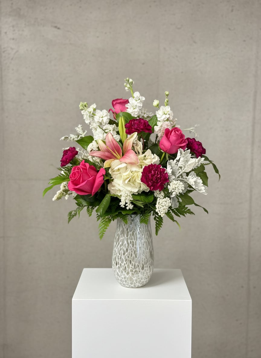 A Mother’s Embrace  - “A Mother’s Embrace” is a tender, heartfelt arrangement designed to feel warm, graceful, and enduring. Set in a hand-blown white iridescent vase that shimmers softly with light, it serves as a beautiful keepsake long after the blooms have faded.  Cloud-like white hydrangeas form a soft foundation, while pink lilies rise elegantly, adding movement and a gentle fragrance. White stock weaves through the design, bringing height and a fresh, airy texture. Hot pink roses provide a vibrant focal point, symbolizing love and appreciation, balanced by the subtle charm of purple carnations.  Dusty miller adds a silvery, velvety softness throughout, and white statice offers delicate accents that tie the palette together. Designed all around, the arrangement feels complete and nurturing from every angle—like a warm, floral embrace meant to honor and celebrate mom.