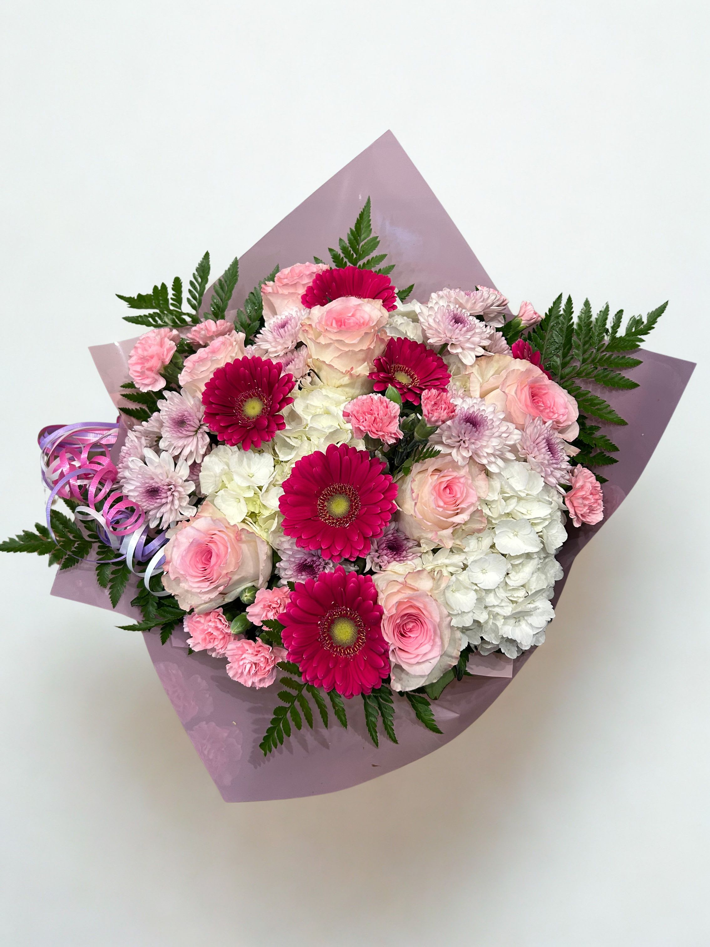 Pretty in Pink - Gerbera daisies pop against a bed of pale pink roses and creamy white hydrangeas in this cheerful birthday bouquet-an ideal birthday, anniversary, or Mother's Day gift that reads romantic and celebratory at once.  This full, rounded arrangement also includes soft pink carnations and small lavender chrysanthemums for texture and movement, all wrapped in lavender wrapping paper with a curled decorative ribbon for a polished, gift-ready presentation. 