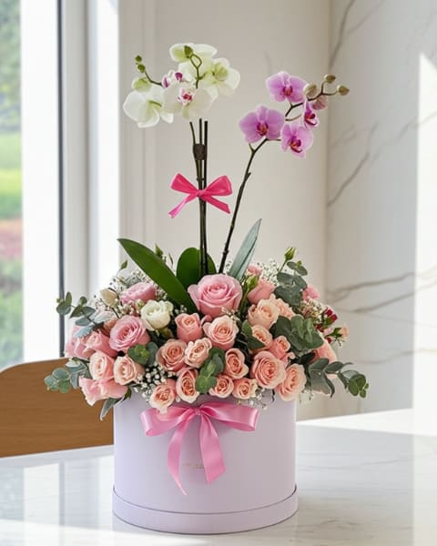 Enchanted Garden Bloom Box - A dense cluster of pink and peach roses paired with two arching Phalaenopsis orchid stems gives this flower arrangement a romantic, elegant look perfect for an anniversary bouquet, birthday gift, or a heartfelt thank-you. The white and lavender tones of the orchids rise above the soft pastel roses to create graceful height and a sculpted silhouette that will make the recipient feel genuinely celebrated.  Arranged tightly in a round lavender hat box and tied with a bright pink ribbon, the presentation reads polished and gift-ready-the kind of boxed floral gift that feels special the moment it's set down. Colors include pink, white, purple, and fresh green foliage; the orchids add a silky texture while the roses bring plush, classic bloom shapes.  A thoughtful choice for the person you want to honor with something both delicate and refined, this keeps well with standard cut-flower care: keep water fresh, place away from direct sun and drafts, and rehydrate the foam or liner as needed. The compact hat-box size makes it easy to display on a table, desk, or bedside-an elegant surprise that says you were thinking of them.