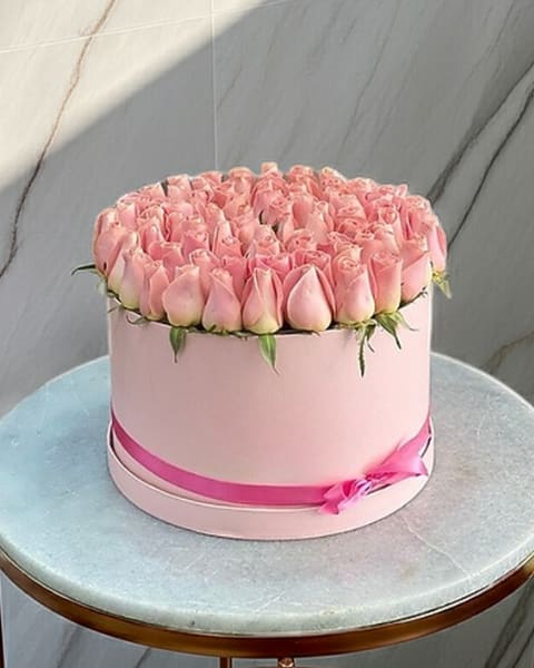 Pink Blush Bloom Box - A lush dome of pale pink roses makes an immediate, romantic statement-perfect as an anniversary bouquet, birthday surprise, or Mother's Day gift. The Pink Blush Bloom Box is a compact flower arrangement that reads soft and polished from every angle, designed to make the person you're thinking of feel cherished the moment they see it.  Tightly packed pale pink roses form a dense, rounded cluster set into a round blush hat box, the monochrome palette softened by small touches of green at the base for a natural, garden-fresh contrast. A bright pink ribbon tied around the box adds a feminine, celebratory flourish while keeping the presentation neat and refined. The low, compact shape makes it ideal for tabletops, bedside tables, or a desk without overwhelming the space.  This arrangement is made to celebrate someone special-mother, partner, friend-who deserves to feel seen and loved. The compact size is easy to display; refresh water and trim stems if repotted, or enjoy the finished boxed presentation as an elegant, no-fuss gift.