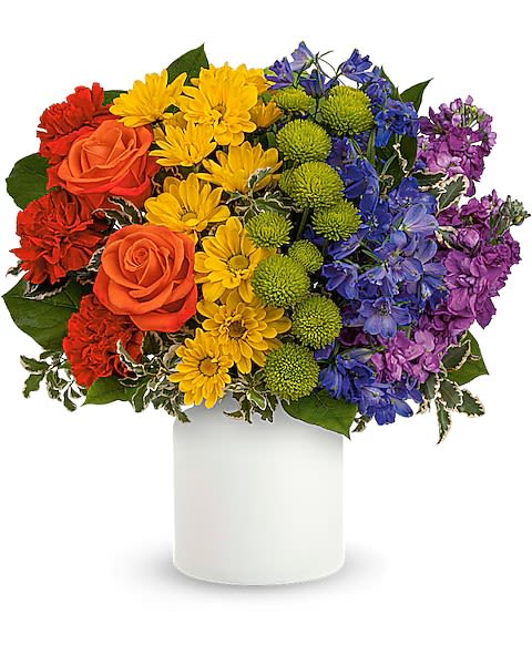 Rainbow Love - A bold, joy-filled arrangement that reads like a pocket-sized rainbow - perfect as a birthday bouquet, a cheerful thank-you, or an everyday gift to brighten someone's day. This mixed-color flower arrangement places the recipient first: the layered bursts of red and orange roses, vivid yellow daisy-like blooms, and pops of purple and blue make the moment feel festive and unmistakably celebratory.  Compact but full-bodied, the design groups color in bold sections for a vivid visual impact. You'll find classic roses paired with chrysanthemums, daisies, green button mums, and complementary blue and purple filler flowers, all arranged tightly for a rounded, sculpted silhouette. The bouquet arrives in a clean white ceramic vase that highlights the colors and fits easily on a bedside table or desk.  Give this when you want someone to feel genuinely cheered and seen - whether marking a birthday, saying thanks, or just because. The sturdy ceramic vase makes it an easy-to-display keepsake; refresh water and trim stems as needed to extend bloom life.