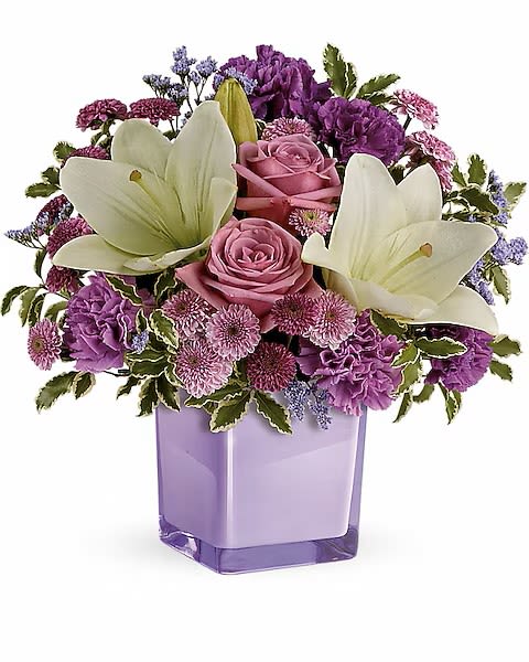 Pleasing Purple - Two large white lilies open at the top of this rounded flower arrangement, their perfume and pure petals brightening a cascade of pink roses and dense purple and lavender blooms-perfect as a birthday bouquet, thank-you gift, or everyday arrangement to make someone feel cherished. The overall mood reads romantic and cheerful, designed to make the recipient feel genuinely seen and celebrated.  Pleasing Purple pairs soft pink roses with carnations, chrysanthemums and asters in varying purple tones for a layered, full-bodied silhouette; flowers are packed closely for a lush, abundant look. The stems sit in a lavender glass vase that reinforces the palette and gives the gift a polished, ready-to-display finish.  This arrangement suits a friend, partner, or colleague who appreciates soft color contrasts and a fragrant focal bloom; it arrives ready to enjoy and brightens a bedside table, dining room or office. Care is simple-keep water fresh and trim stems if needed to extend vase life.