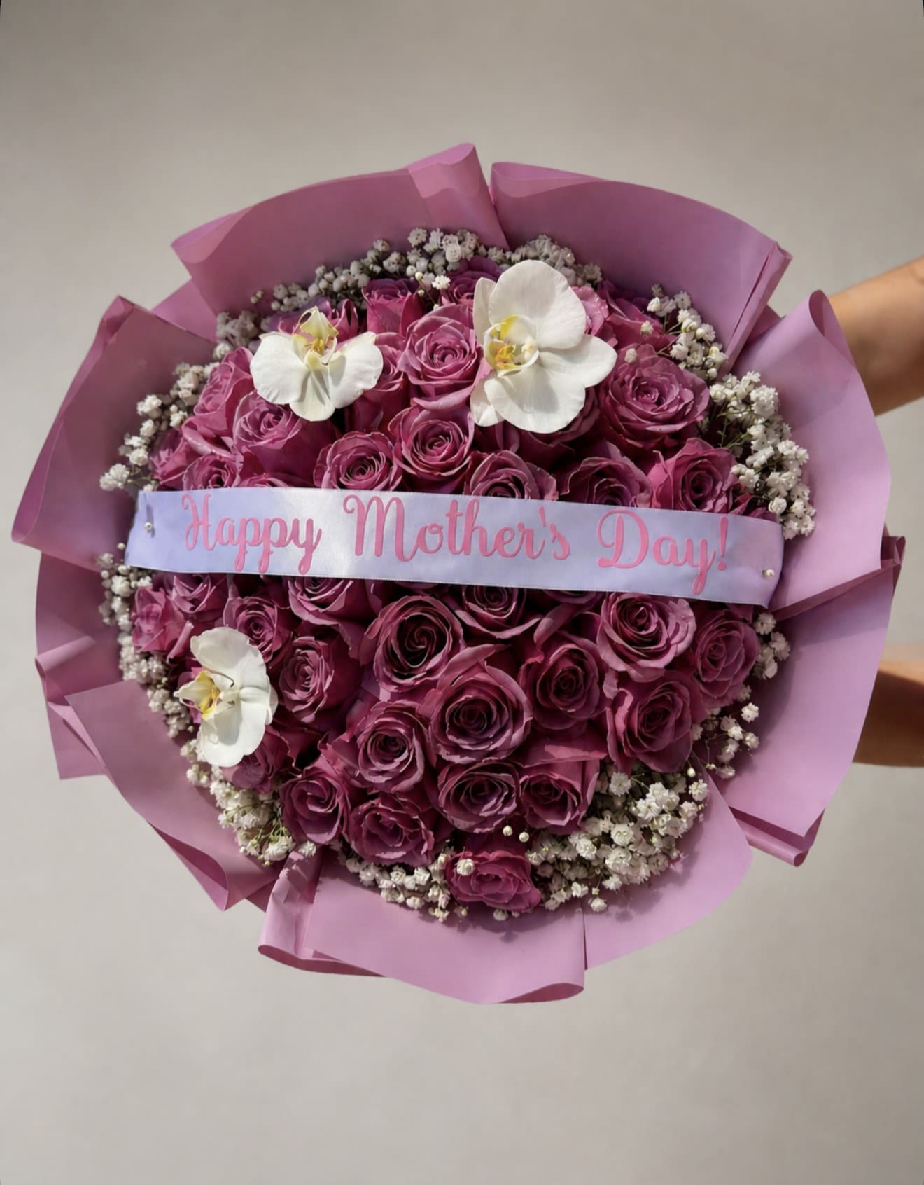 Lavender Mom  - A lush, round Mother's Day flower arrangement that reads like a love letter in pinks and purples-perfect as a birthday bouquet or a springtime gift for mom. The dense cluster of vivid roses creates immediate visual warmth, while three pure white orchids lend an elegant, unexpected focal point; the included ribbon banner saying "Happy Mother's Day!" makes the sentiment unmistakable and affectionate.  This arrangement pairs soft lavender paper wrap with a delicate ring of tiny white filler flowers for a textured, romantic silhouette. Colors are mainly purple, white, and pink; flowers are roses and orchids; finished with a decorative ribbon banner that reinforces the celebratory message. The compact, round shape is designed to sit beautifully on a kitchen table or bedside-an arrangement that will make the person you're thinking of feel seen and cherished.  Ideal for Mother's Day, a spring anniversary, or to say "thank you," Lavender Mom arrives ready to gift and display. For best enjoyment, place in fresh water on arrival and recut stems if you transfer them to a vase; order in time for holiday delivery to ensure it arrives at peak bloom.