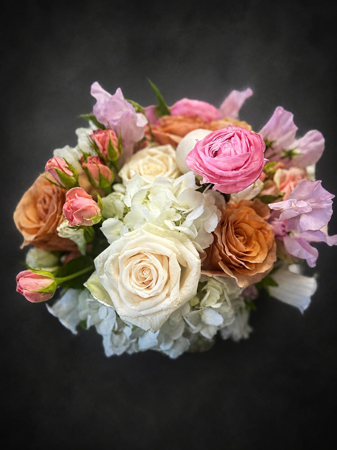 A Mother’s Love - Soft white and blush roses crown this compact arrangement, a rounded bouquet called for celebrating Mom or marking a quiet anniversary. The white blooms brighten the center while peach-toned roses add warm depth, and the overall effect reads as a graceful, feminine gift-perfect for Mother's Day, a thank-you, or a sweet romantic gesture.  Pale hydrangea clusters fill and soften the shape, while soft pink and lavender sweet pea trails lend an airy, garden-style texture around the edges. The mix of white, pink, peach and purple creates a gentle, layered palette that feels both elegant and approachable; this is the kind of bouquet that makes the person you're thinking of feel genuinely seen and cherished.  Sized as a compact, rounded arrangement, it fits comfortably on a bedside table or dining centerpiece and arrives ready to display. For best enjoyment, keep blooms in fresh water away from direct heat-replace water and recut stems every few days to prolong their beauty.