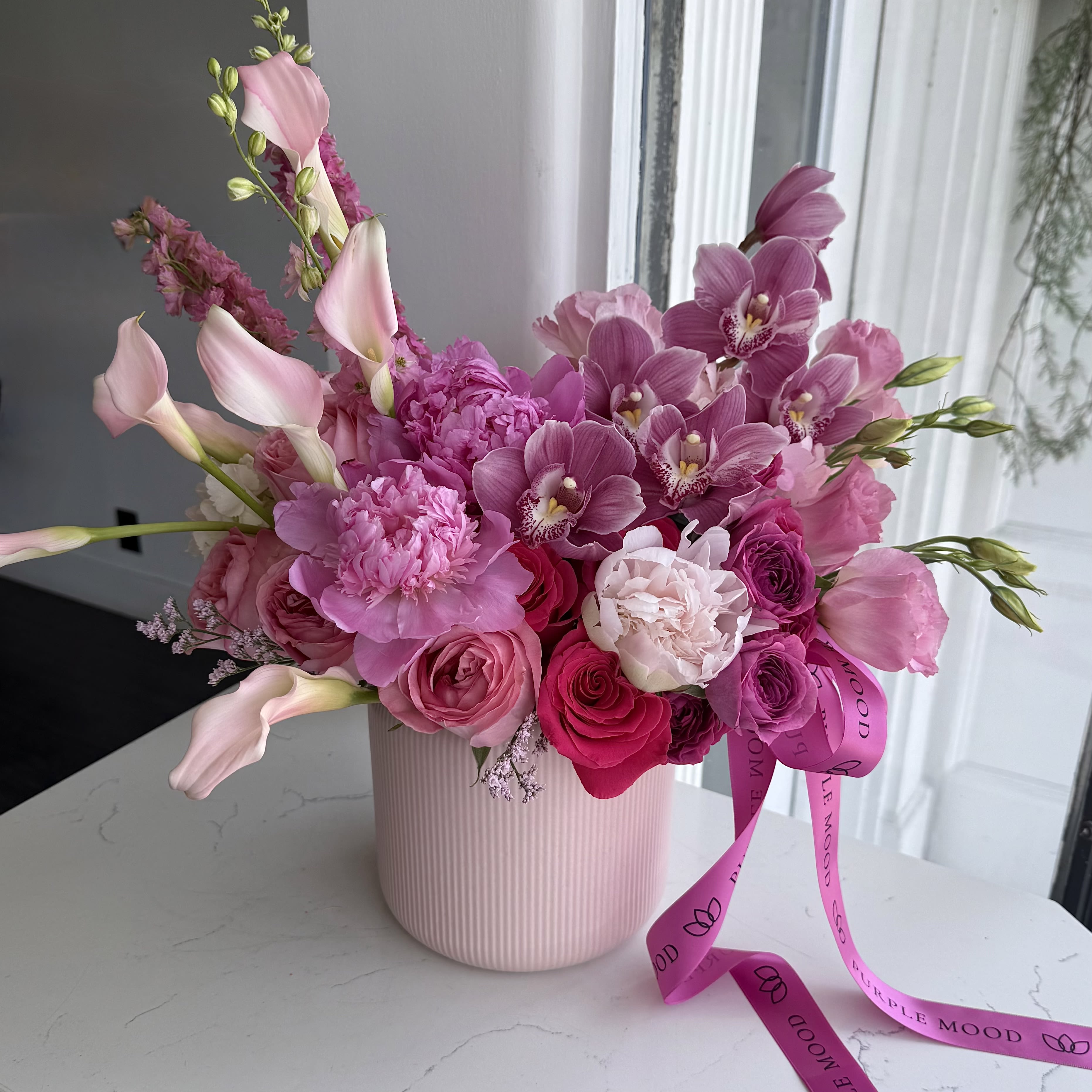 Pink Noir Vase - Soft symphony in pink and purple. Graceful calla lilies dance alongside rich orchids, complemented by delicate roses and fluffy peonies. Nestled in a gentle pink ribbed vase and finished with a flowing satin ribbon, this bouquet feels both tender and quietly seductive. Timeless elegance with a modern romantic soul.