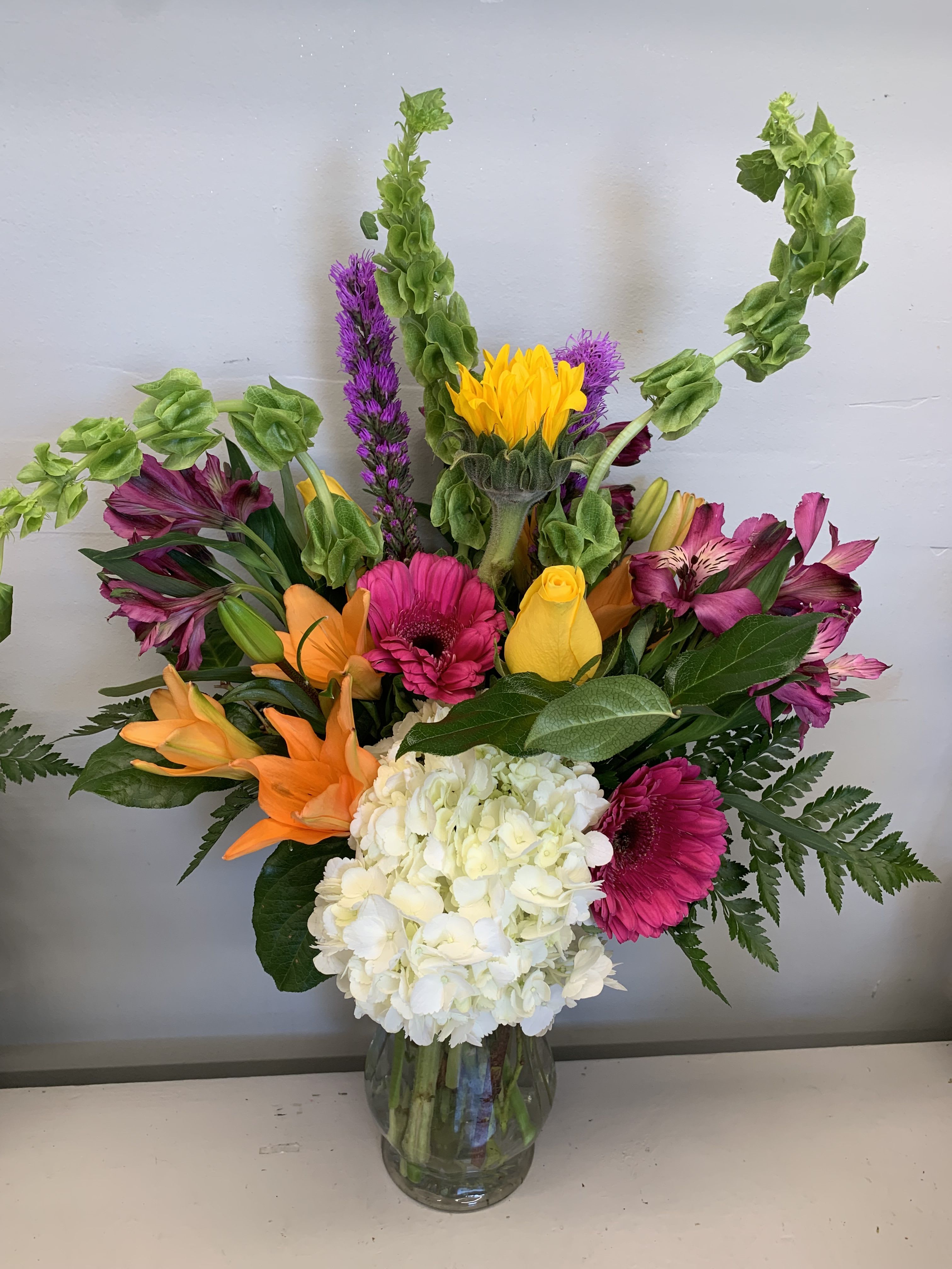 Tall mixed bouquet with white hydrangea, orange lilies, pink gerbera, yellow blooms and green Bells of Ireland in a clear vase