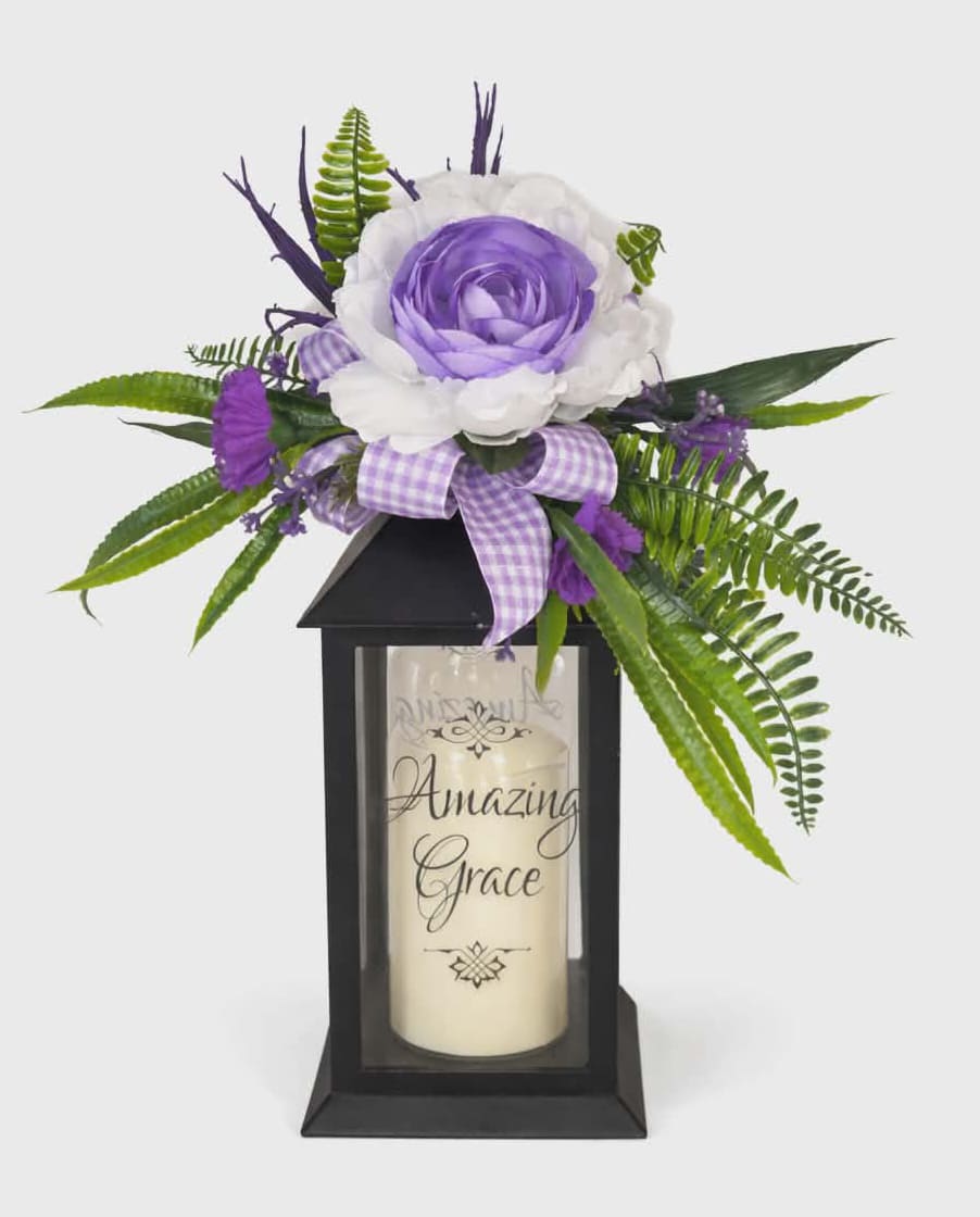 "Amazing Grace" Lighted Lantern - A soft glowing lantern topped with a silk rose creates a comforting focal point for sympathy, a thoughtful get well gift, or a peaceful housewarming accent. This lighted lantern arrangement pairs a cream pillar candle printed with "Amazing Grace" and a warm LED glow with a layered cluster of white and lavender silk roses, small purple blooms, and fern-like greenery - perfect for Easter mantels or a gentle "just because" surprise that makes someone feel seen and comforted.  The black rectangular lantern measures 13" high x 5.5" wide x 5.5" deep and includes a flameless LED pillar candle with a 6-hour on / 18-hour off automatic timer (3 AAA batteries included). A lavender-and-white gingham ribbon bow nests among the flowers for a homespun touch, and the weather-friendly construction allows hanging or free-standing display on a table, shelf, or mantel. Ideal for birthdays, Passover, sympathy and funeral remembrances, or as a serene accent when welcoming someone into a new home.