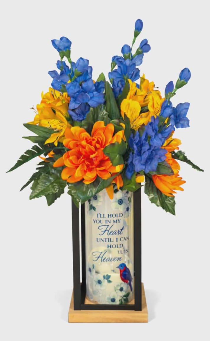 "Hold You In My Heart" Silk Cylinder Lighted Lantern - Silk vivid blue delphinium and bright orange chrysanthemums cluster with sunny yellow and soft white alstroemeria above a tall glass cylinder, creating a lantern-style sympathy arrangement that gently brightens a memorial space. This silk flower arrangement for sympathy and funeral display pairs bold, lasting color with a comforting message-perfect for the person you're remembering or the family you wish to support.  The piece is built into a black metal lantern frame on a wooden base, with a printed glass cylinder featuring small blue blossoms, a colorful bird illustration, and the remembrance text, "I'll hold you in my heart until I can hold you in Heaven." A flameless LED candle sits inside the cylinder; it runs on an automatic 6-hour ON / 18-hour OFF timer and uses three AAA batteries (included). Dimensions: 4.75" W x 8.5" H x 4.75" D. Designed for indoor display, it offers a tasteful, long-lasting tribute that will make the recipient feel seen and comforted.