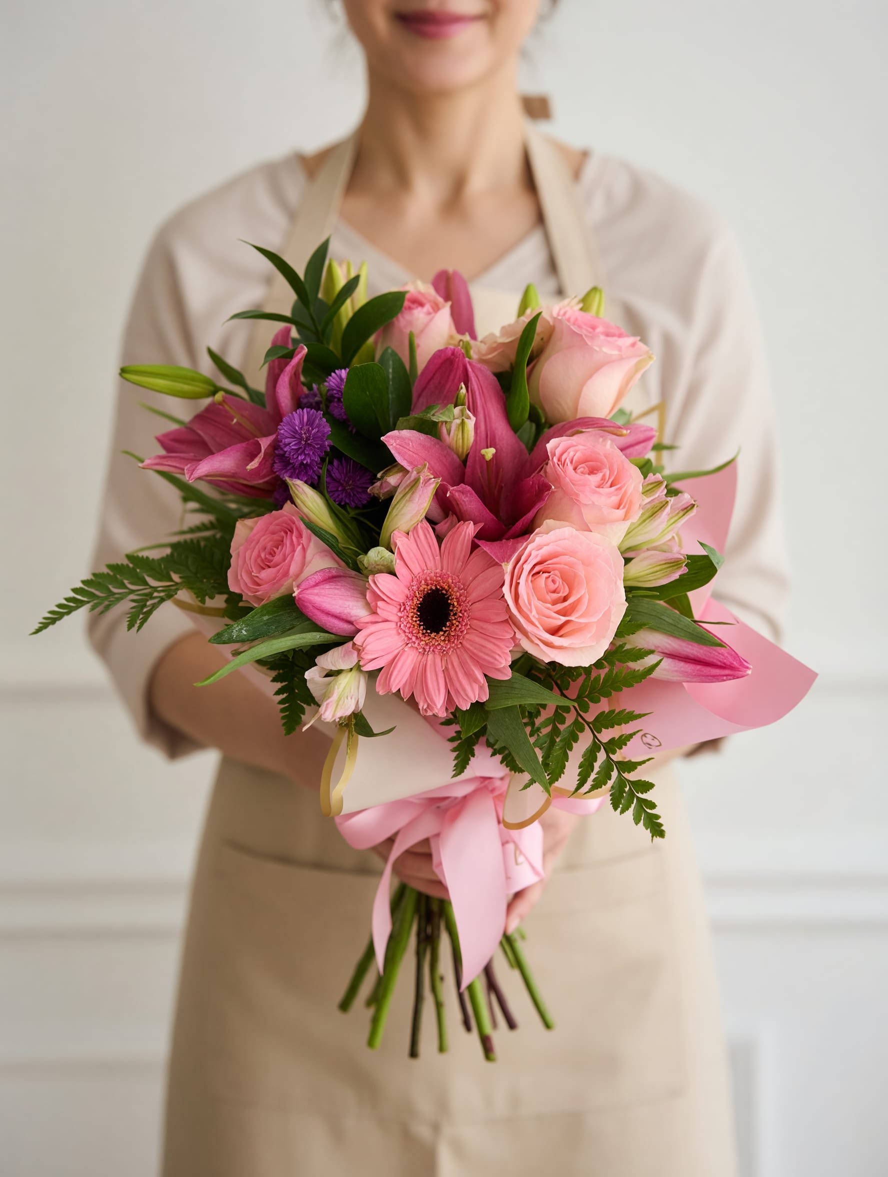 Pink whisper  - Soft pink roses, a single pink gerbera daisy and bold magenta lilies mingle with purple asters and delicate alstroemeria in this hand-tied flower arrangement-perfect as a birthday bouquet, anniversary surprise, Mother's Day gift, or a cheerful thank-you. The blush, fuchsia and violet tones read romantic and joyful, so the person you're thinking of will feel genuinely celebrated the moment they see it.  This full, rounded bouquet uses layered petals and visible lily buds for a fresh, just-arranged look, with greenery for contrast and movement. It arrives wrapped in pale pink paper and tied with a satin ribbon for a polished, gift-ready presentation; soft textures and a springlike color palette make it ideal for everyday gifting or special moments when you want someone to feel truly seen.