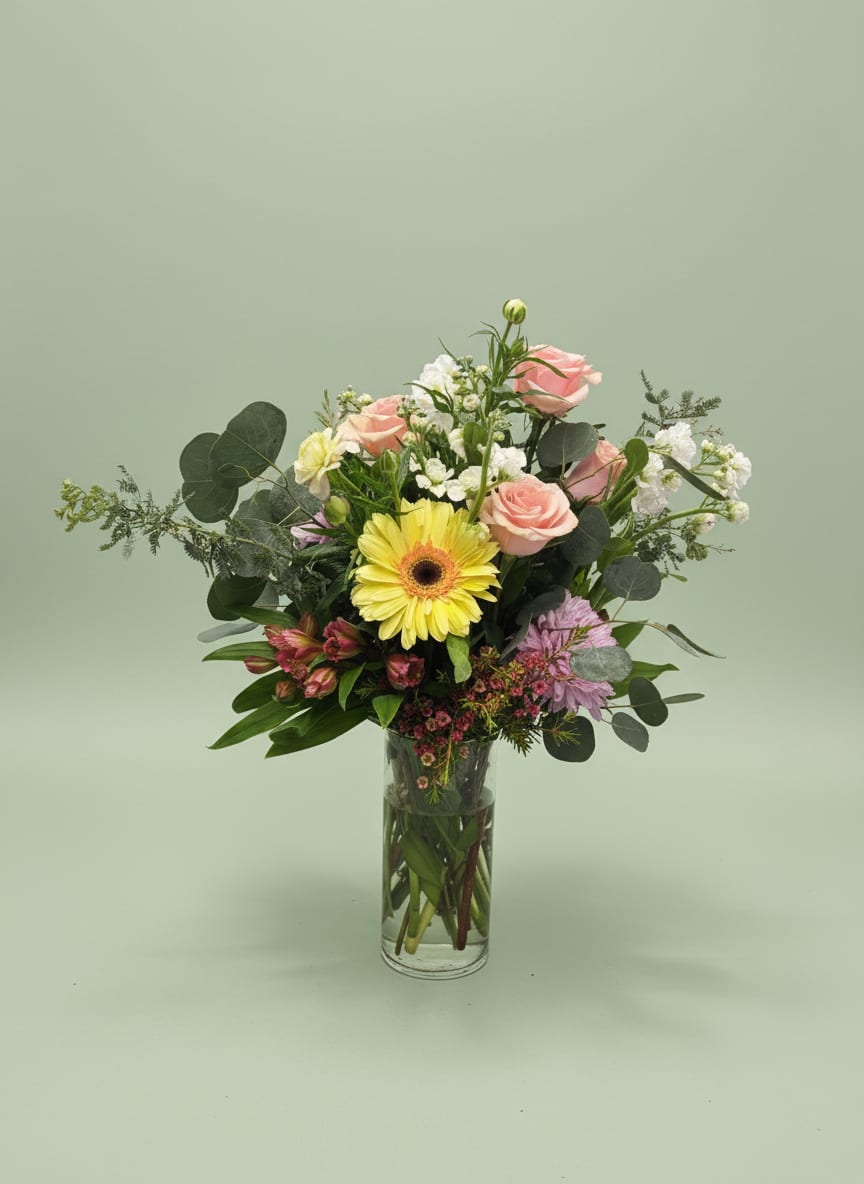 Blush crush  - A single bright yellow gerbera daisy anchors this airy garden-style flower arrangement, making the mixed bouquet feel instantly cheerful - perfect as a birthday bouquet, a thank-you gift, or a spring celebration arrangement. Soft pink roses and pale lavender accents create a romantic, pastel palette that lets the yellow focal flower sing, so the person you're thinking of will feel noticed and genuinely celebrated.  Packed loosely in a tall clear glass vase, the design mixes white clustered blooms, carnations, stock, alstroemeria, chrysanthemums and rounded eucalyptus-like foliage for a fresh, layered texture. The silhouette is asymmetrical and full without being fussy, with fine fillers and visible stems in the cylindrical vase lending a modern, natural look.  This everyday yet special arrangement arrives ready to display and brightens a bedside table, kitchen counter, or office desk. Choose this if you want a light, uplifting bouquet that's easy to care for - replace the water every couple of days and trim stems slightly for the longest enjoyment.