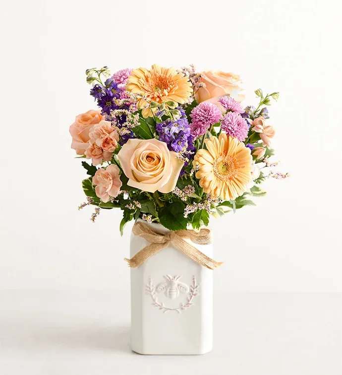 Honey Bee Buzz - Get them buzzing with our charming bouquet! A garden-fresh gathering of blooms in sweet shades of lavender, purple, peach and pink come together for a mix of color and texture. Designed in our Sweet As Can Bee artisanal vase, featuring an adorable honey bee and raffia bow, it’s a gift that adds quaint beauty to any decor.