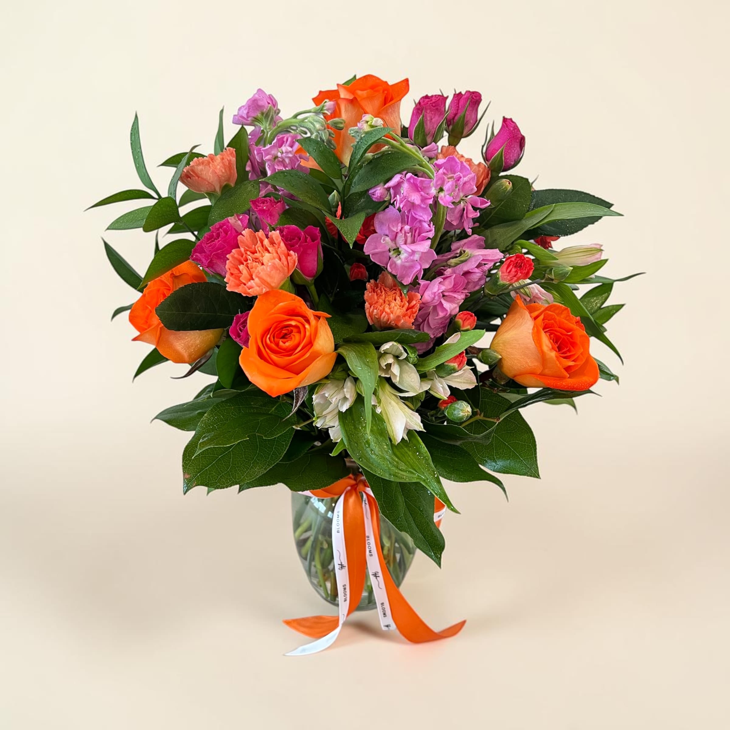 Bright Smiles  - “Bright Smiles” is a cheerful, uplifting arrangement designed to instantly lift spirits and warm hearts. Vibrant orange roses take center stage, symbolizing enthusiasm and joy, while playful pops of pink blooms add a touch of sweetness and charm. Soft accents of carnations and delicate filler flowers create texture and fullness, all nestled in rich, lush greenery for a fresh, garden-inspired feel.  Presented in a clear glass vase and finished with a bright ribbon, this bouquet radiates happiness from every angle. Perfect for celebrating special moments—or simply reminding someone they’re loved—Bright Smiles delivers a burst of sunshine in floral form.