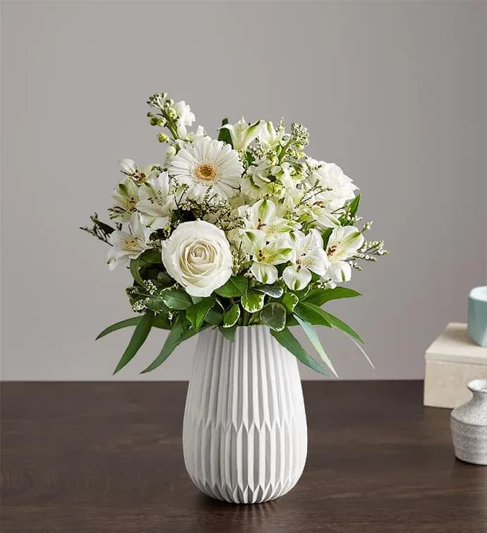 Treasured Memories All White - A serene all-white arrangement designed to comfort and celebrate-the ideal sympathy arrangement, wedding centerpiece, or thank-you gift. Treasured Memories All White pairs classic white roses and lilies with daisy-like gerbera focal blooms and delicate alstroemeria to create a calm, uplifting presence that makes the recipient feel genuinely seen and cared for.