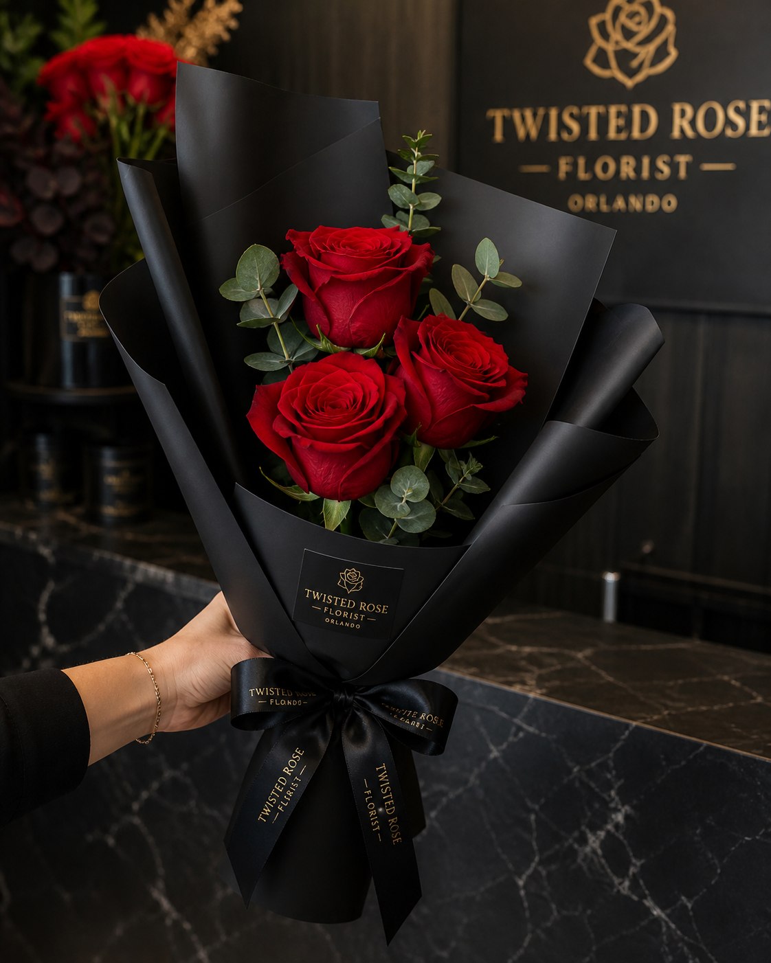 Rose Wrap Special - Three deep red roses set against matte black wrap create an instantly dramatic, romantic gift-perfect as an anniversary bouquet, Valentine's Day surprise, or an elegant everyday gesture for the person you want to make feel truly seen. The compact, hand-held arrangement reads polished and boutique-style; its bold red-and-black palette delivers a formal, upscale impression that says devotion with minimal fuss.  Tightly clustered red roses are framed in black wrapping paper and finished with a black satin ribbon (and in some versions a black ribbon printed in gold), while small eucalyptus sprigs introduce a muted green contrast that highlights the petals' richness. The rounded, hand-tied form is compact and easy to carry, ideal for a personal celebration or a thoughtful drop-by gift. Simple care: keep stems in fresh water, trim ends when arranging, and present within a day or two for best bloom impact.