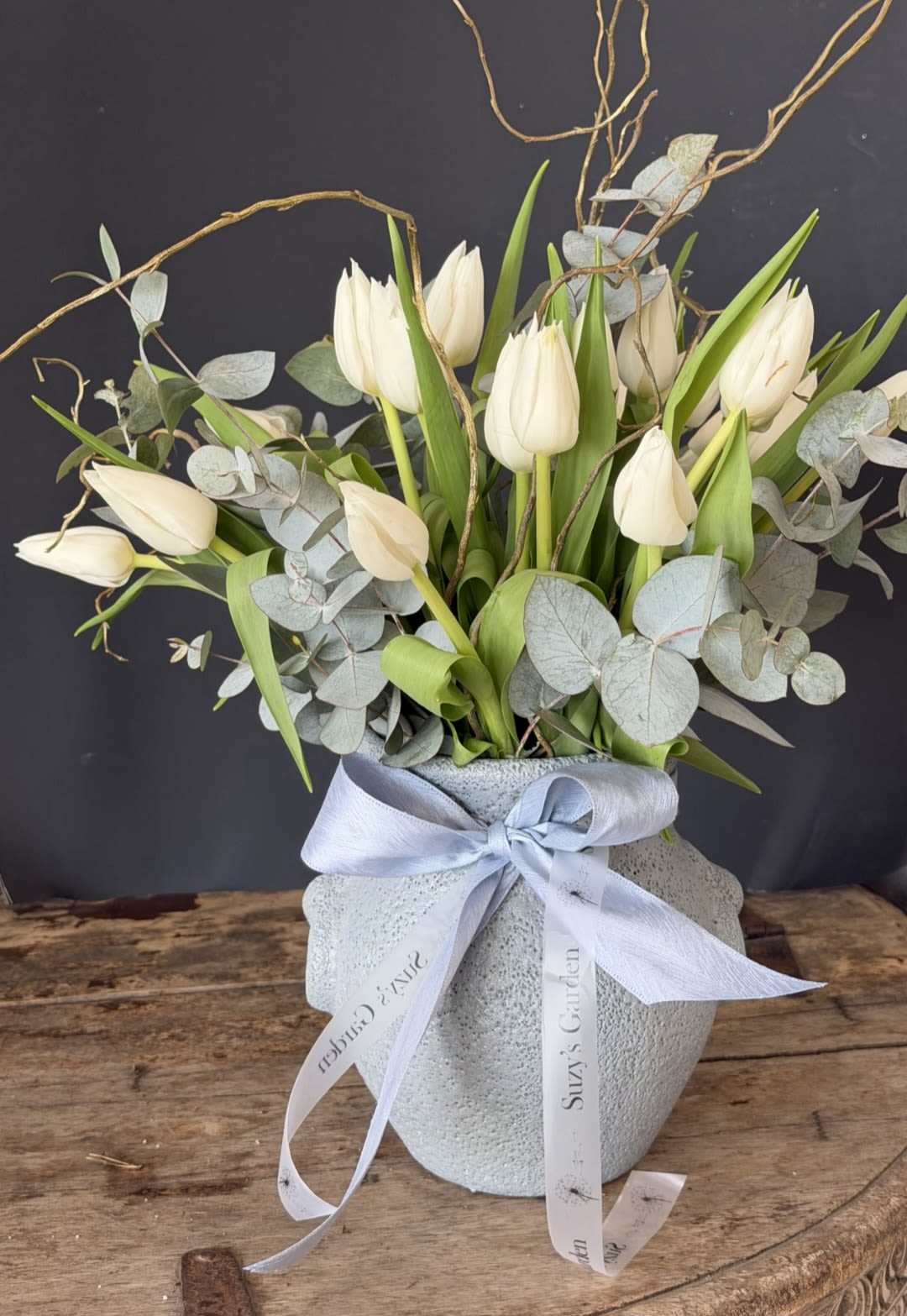 Whisper Tulip Arrangement - Elegant white tulips with soft eucalyptus and natural branches, beautifully arranged in a textured ceramic vase with a delicate ribbon for a calm, refined look.