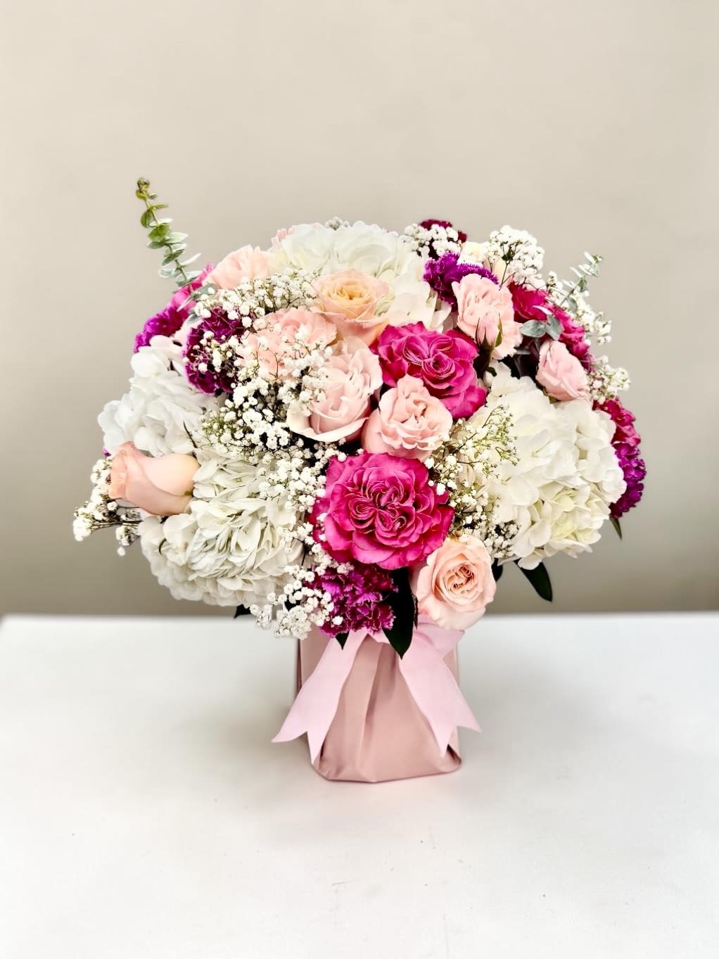 Pink Magenta by Daisy - Lush pink and white roses mingle with voluminous white hydrangeas and delicate sprays of baby's breath for a romantic birthday bouquet or anniversary gift that feels instantly polished. Bright magenta accents pop against the softer tones, giving the arrangement a lively, feminine energy that reads as both celebratory and intimate-perfect for surprising the person you're thinking of on a special day.  Carefully arranged in a blush wrapped vase and finished with a soft satin ribbon, this rounded arrangement blends roses, hydrangeas, carnations, dianthus, and other mixed blooms into a dense, garden-fresh composition. The mix of textures-from the creamy hydrangea clusters to the classic rose petals and tiny baby's breath-creates visual depth and a soft, fragrant presence that fills a room without overwhelming it.  Designed to arrive gift-ready, this bouquet is ideal for anniversaries, birthdays, weddings, or as a thoughtful "just because" surprise that makes someone feel truly celebrated. Order with standard care instructions and the arrangement will stay vibrant for days when kept in fresh water and out of direct heat-an elegant, ready-to-give option for memorable moments.