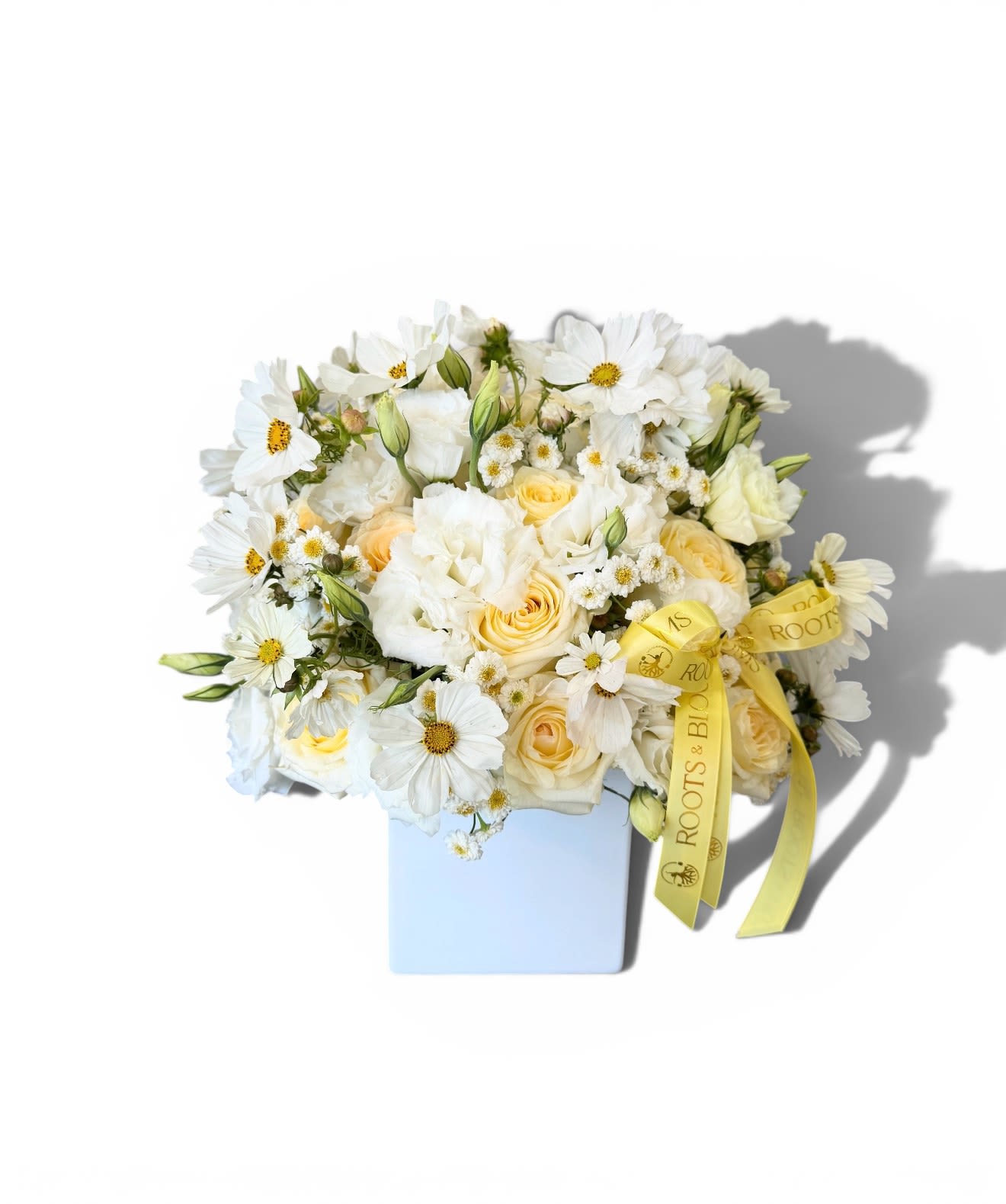 Ivory Sunshine - A dreamy blend of ivory roses, ruffled white blooms, and delicate daisy accents arranged in a modern white vase. Soft, airy, and timeless. -this design brings a fresh, romantic feel with subtle ouches of warmth and charm.