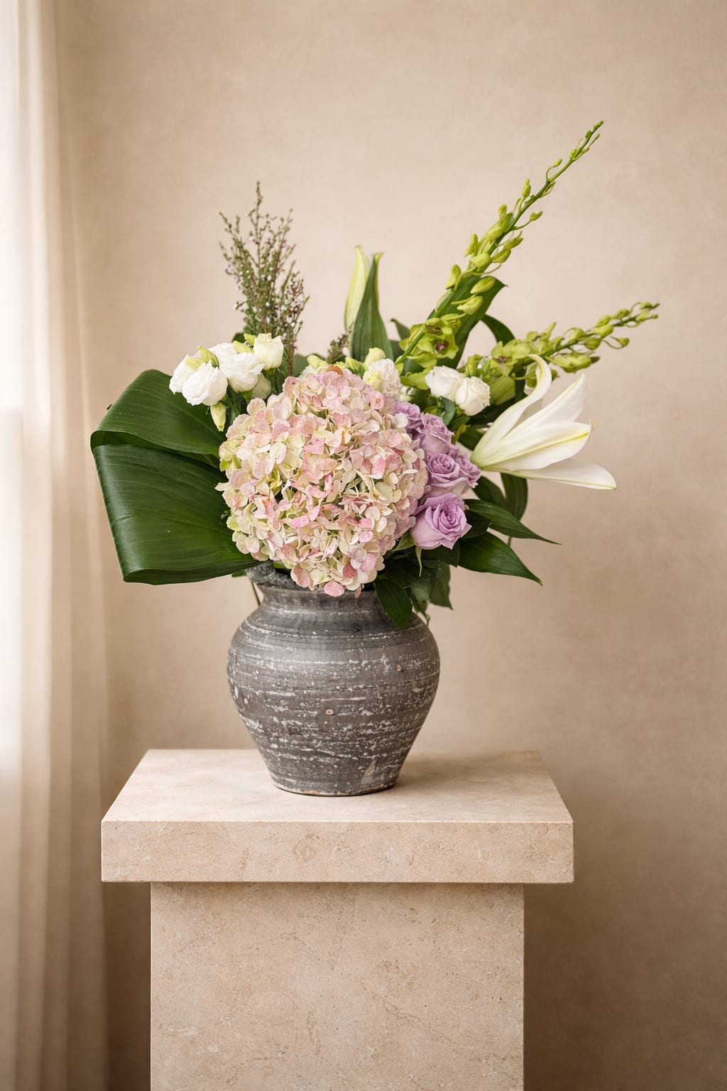 Asymmetrical arrangement of blush hydrangea, lavender roses, white lilies, and broad green leaves in a gray ceramic vase