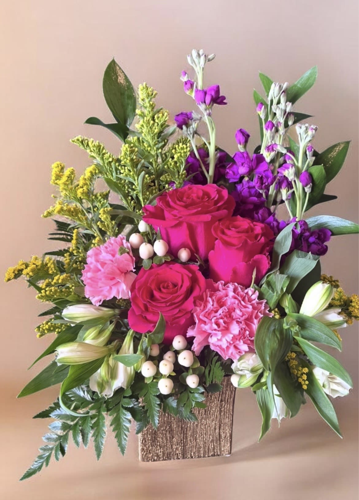 Sweeter Than Ever  - A lush cascade of vivid pink roses and ruffled pink carnations greets the eye-this flower arrangement is a cheerful, romantic choice for a birthday bouquet, anniversary flowers, or Mother's Day gift. The compact rectangular vase and white berry accents give the composition a polished, modern feel while the bright yellow filler and purple clusters lift the palette so the person you're celebrating will feel genuinely noticed and adored.  Soft alstroemeria and delicate white accent blooms mingle with broad green foliage to create a full, layered shape that reads abundant without being fussy. The textured rectangular vase keeps stems neatly contained for a contemporary display; the arrangement's compact proportions make it perfect for tabletops, desks, or a kitchen island where its scent and color can be enjoyed up close.  Send this when you want to say "you're sweet and cherished" - it's ideal for moms, partners, or close friends marking a special day or just because. Care is simple: keep water fresh, trim stems every few days, and place away from direct heat to prolong blooms.