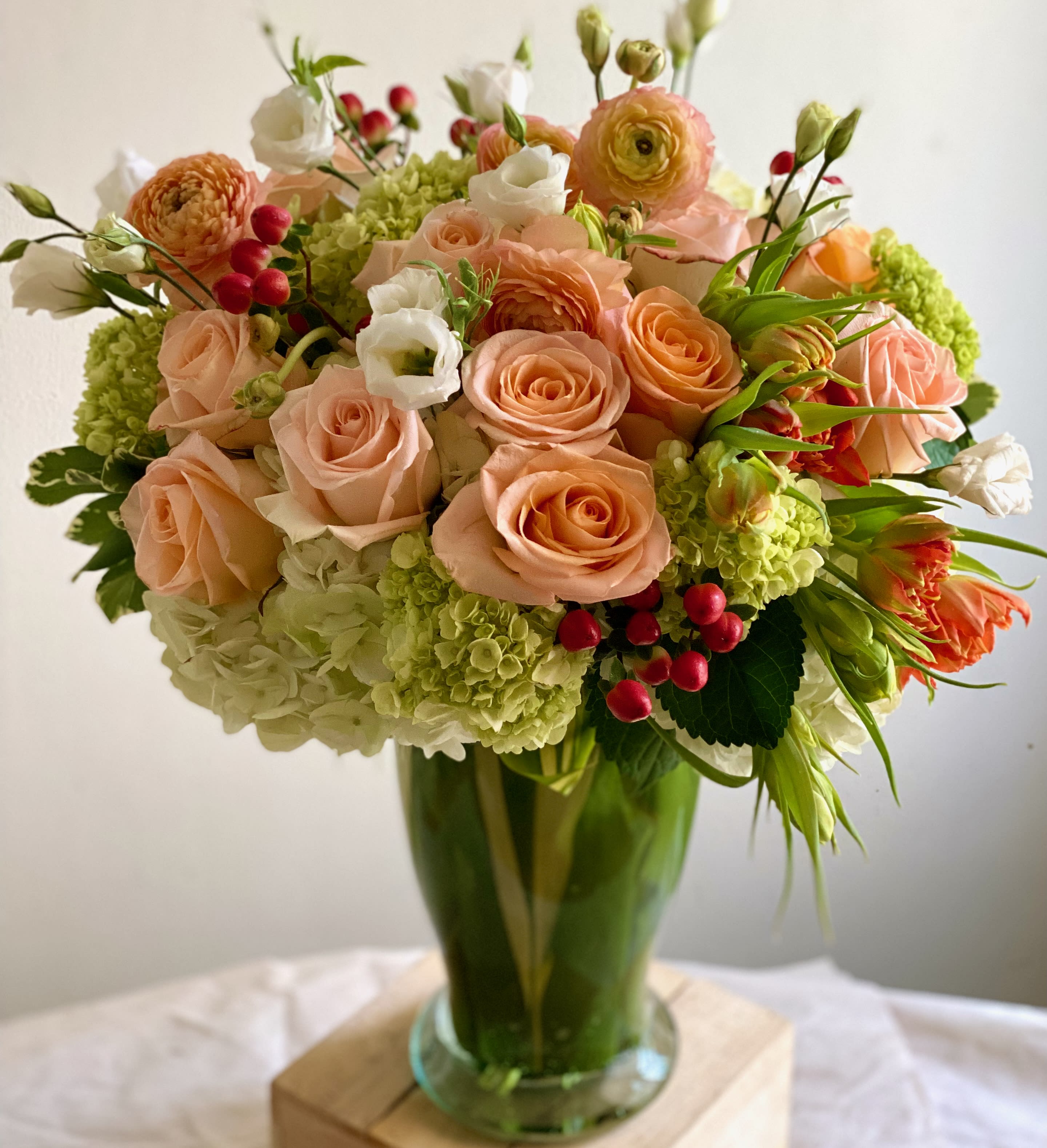 Tall vase arrangement of peach roses, green hydrangeas, white blooms and red berries.