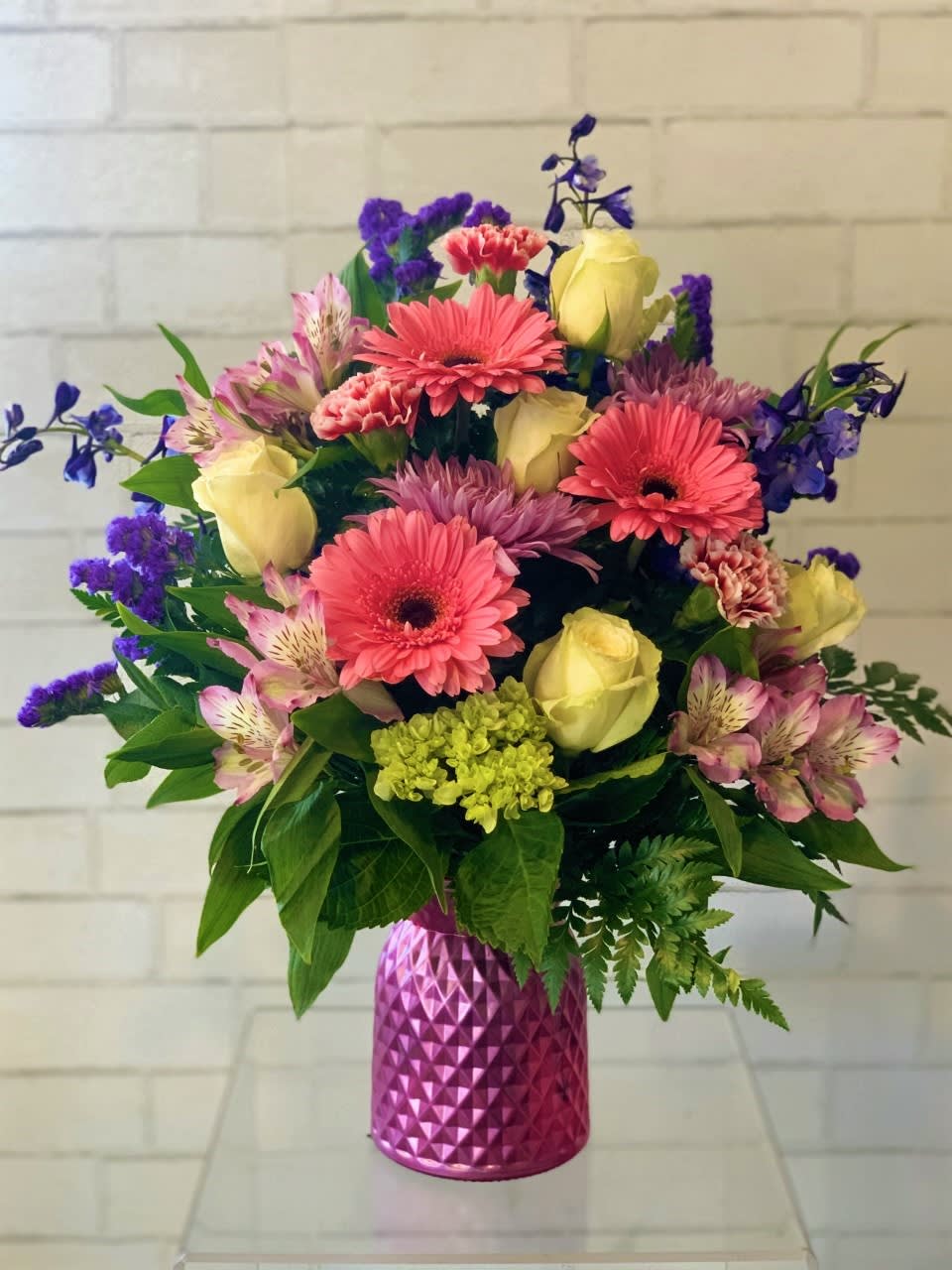 Mixed bouquet of pink gerbera daisies, yellow roses, and purple flowers in a pink vase