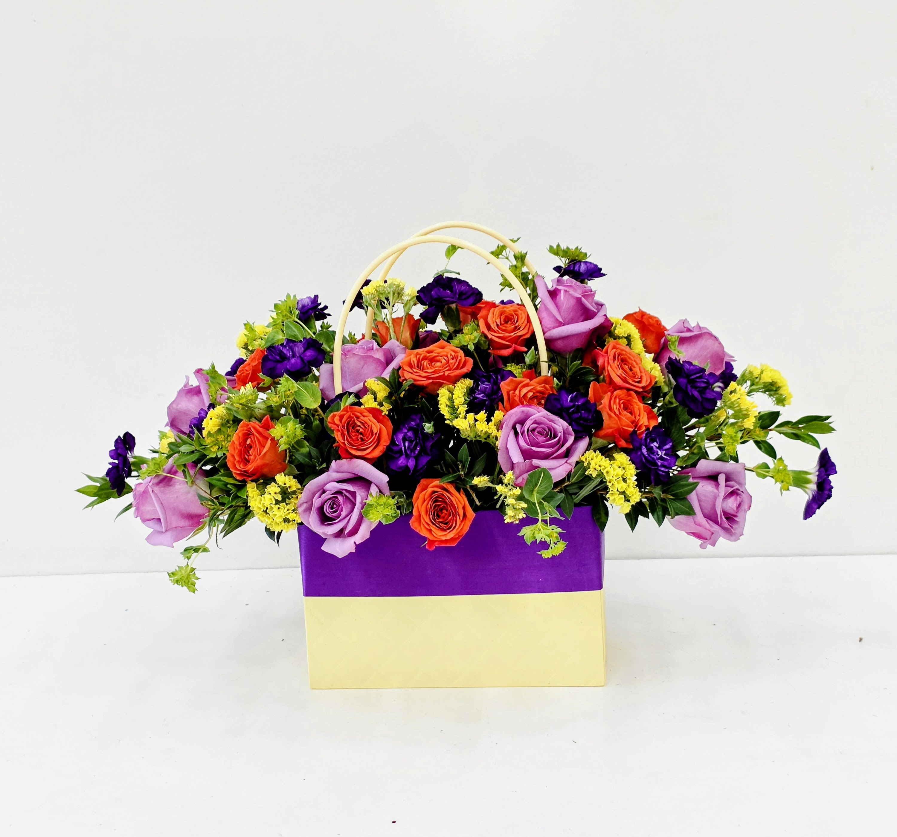 Vivid Blooms in a Bag - Lavender and orange roses mingle with deep purple accents and tiny yellow filler blooms in a playful, celebratory gift arrangement-perfect as a birthday bouquet, congratulations gift, or a cheerful "just because" surprise. The rectangular gift bag presentation with spilling fresh flowers gives the arrangement a casual, modern feel that reads as joyful and abundant; the color contrast makes the person you're thinking of feel noticed and celebrated the moment they see it.  