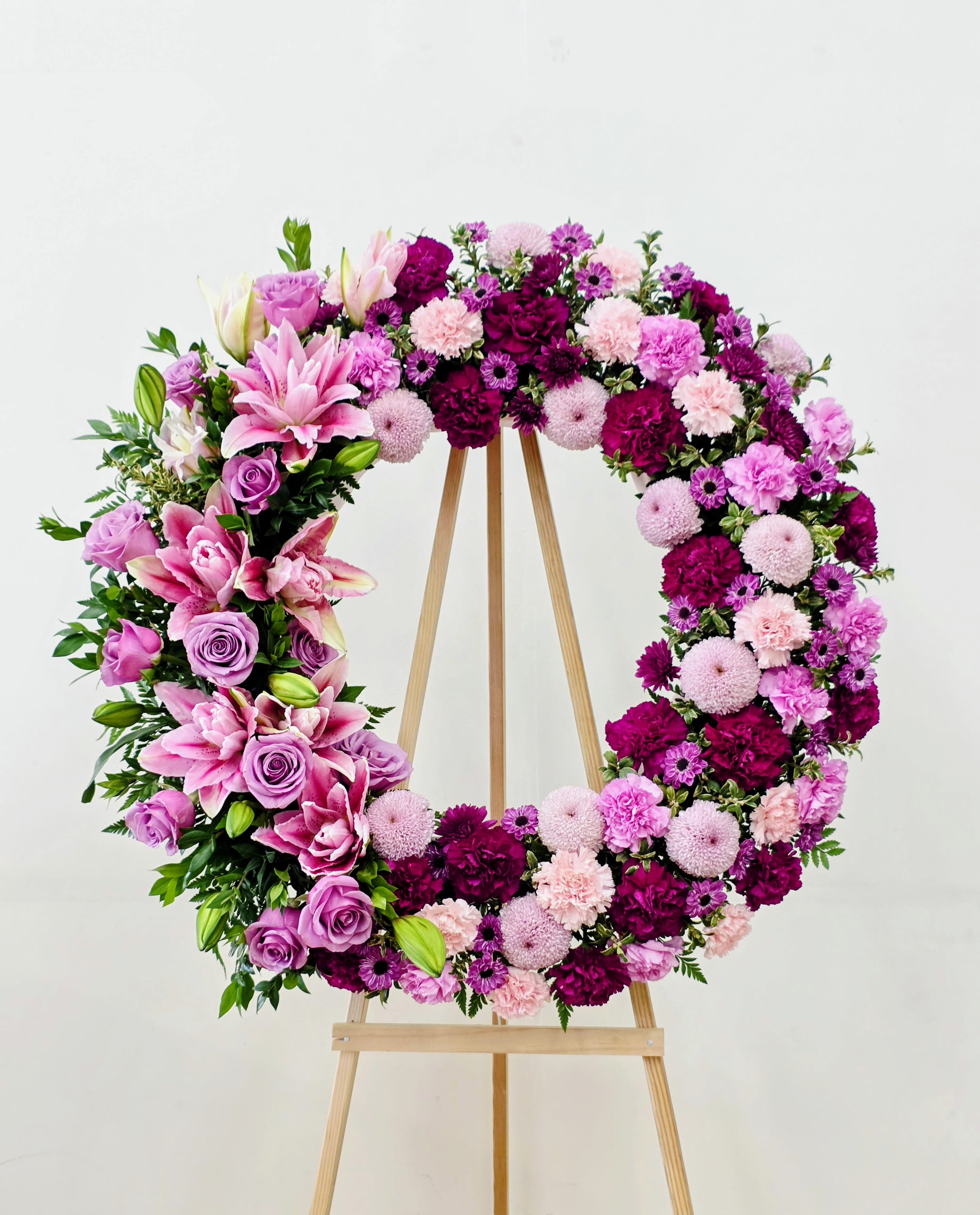 Comforting Wreath - A lush circular wreath in shades of pink, purple, white, and green makes a graceful statement for sympathy, memorial services, or any ceremonial tribute. This standing wreath on a wooden easel presents a solemn-yet-romantic ring of flowers that reads both dignified and comforting-the kind of memorial that helps the person you're honoring feel remembered and cherished.  Choose this wreath when you want to convey sympathy, love, and respect-an arrangement that comforts mourners and honors a life with quiet elegance. Sized for prominent display, it arrives ready on its wooden easel for immediate placement; no special care is needed beyond keeping it sheltered from extreme heat or wind during services.