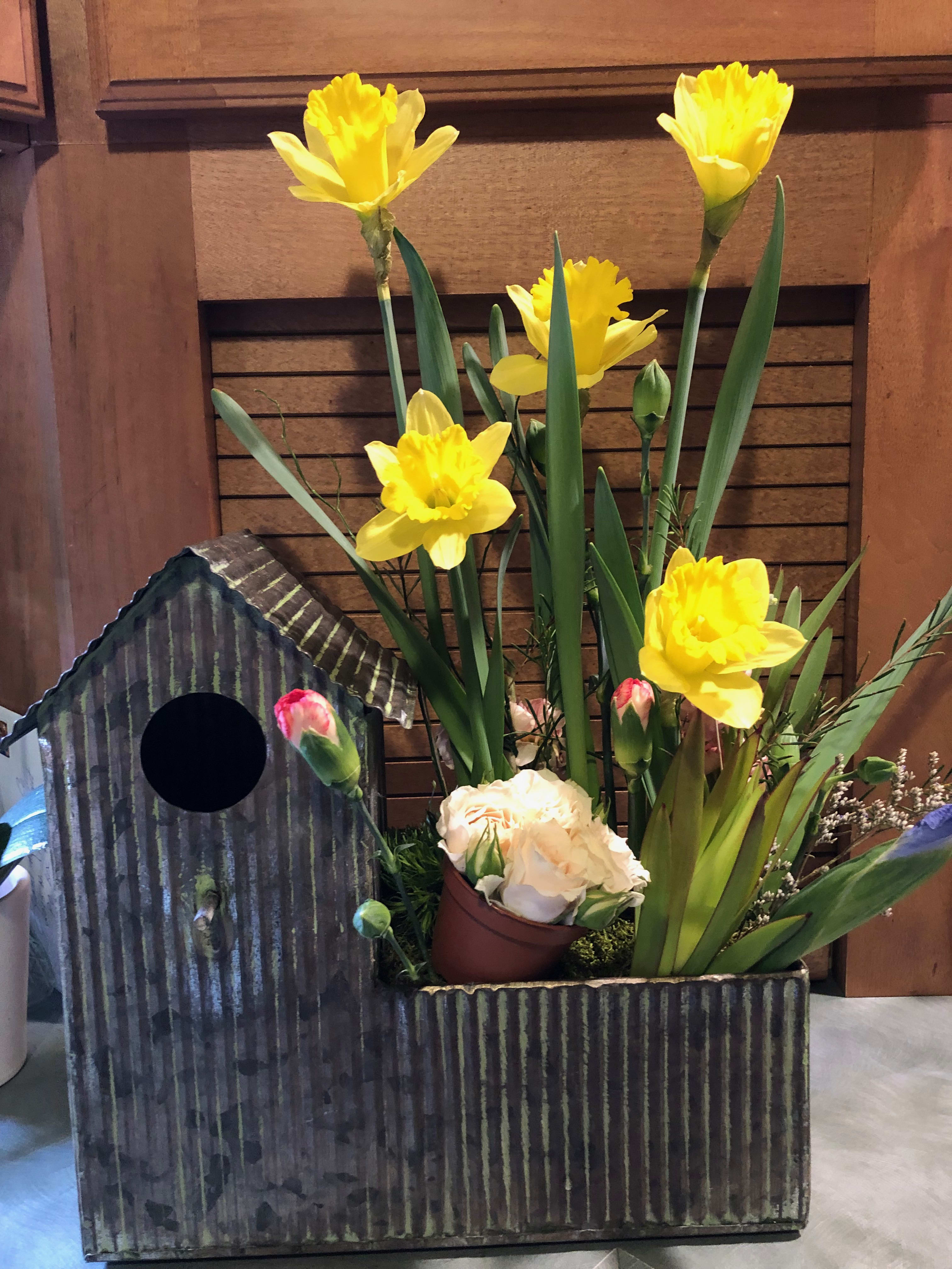Yellow daffodils arranged in a rustic metal container with a small birdhouse accent.