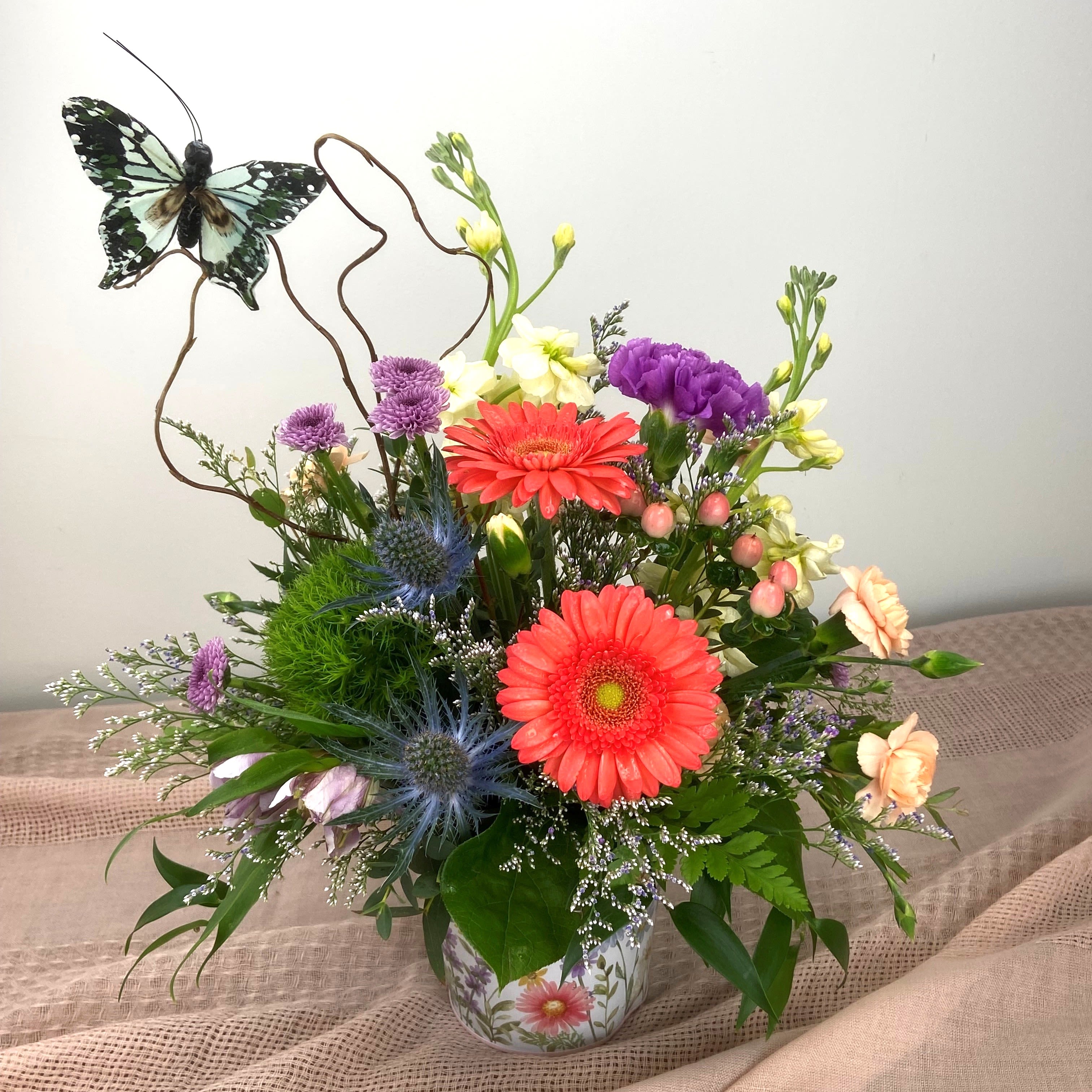Pastels for Mom - Bright coral gerbera daisies take center stage in this pastel garden-style flower arrangement for Mother's Day, birthdays, or a cheerful "just because" surprise. The mix of pinks, purples, whites, greens and soft yellows reads youthful and uplifting, and the whimsical butterfly pick rising above the blooms makes the person you're thinking of feel noticed and celebrated. 