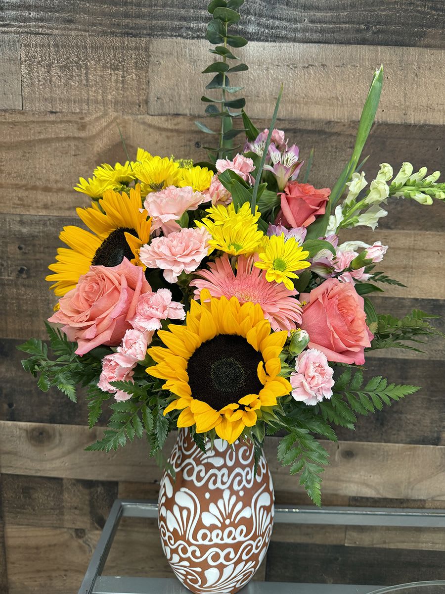 Pretty Sky Mix - Bright yellow sunflowers steal the show in this cheerful mixed bouquet, paired with coral-pink roses and pale pink carnations for a sunny, uplifting birthday bouquet or "just because" surprise. The loose, garden-style shape-with a few taller snapdragons and alstroemeria stems rising above the cluster-creates a joyful, spirited look that will make the person you're thinking of feel noticed and celebrated.  This arrangement mixes yellow, pink, white, and green blooms including sunflowers, roses, carnations, alstroemeria, snapdragons and small yellow daisy-like flowers, arranged in a full, rounded composition with some taller accents for movement. Presented in a patterned brown-and-white ceramic vase, the handcrafted ceramic adds texture and a decorative touch while keeping the focus on the vivid flowers. Ideal for everyday gifting, birthdays, or to brighten someone's day; vase-arranged for easy display and simple care-change the water every few days and trim stems to help the bouquet last.
