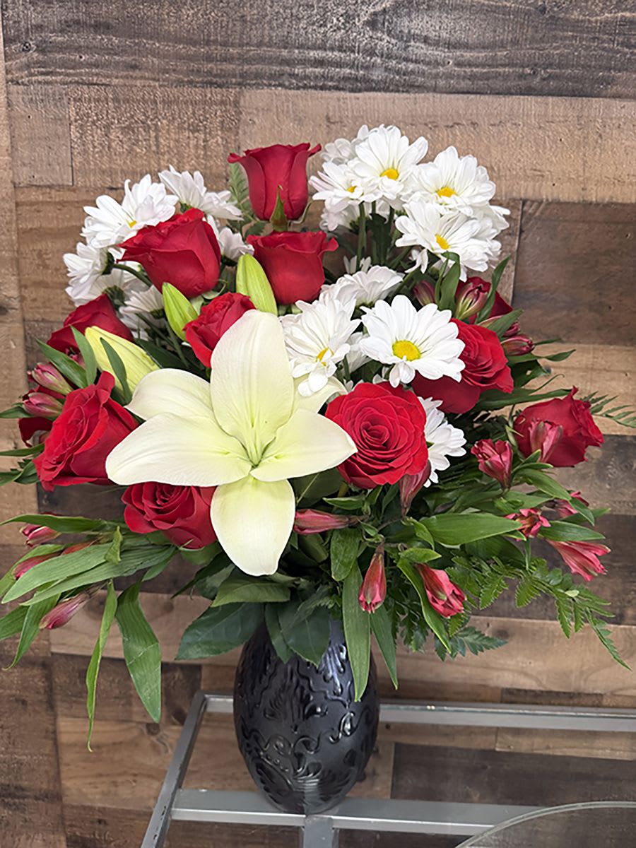 Black Diamond - A striking contrast of deep red roses and crisp white blooms makes this flower arrangement an unforgettable choice for an anniversary, Valentine's Day, or to say congratulations. The "Black Diamond" arrangement centers a showy white lily amid clusters of white daisies and rich red roses, creating a bold red-and-white palette that reads romantic and celebratory-perfect for the person you want to make feel truly seen.  The design is full and rounded, with mixed greenery and delicate pink alstroemeria buds woven around the edges for texture and soft color variation. Presented in a decorative black vase, the dark base adds visual weight and modern elegance while highlighting the vivid reds and snowy whites. The mix of roses, lilies, daisies, and alstroemeria gives varied shapes and scents that feel luxurious without being fussy.  This is the kind of arrangement that works equally well as a romantic gesture, a formal congratulatory gift, or a centerpiece for special moments-she'll feel celebrated the minute it arrives. Order with confidence for same-week delivery when available; keep stems in fresh water and trim a bit every few days to extend the life of the flowers.