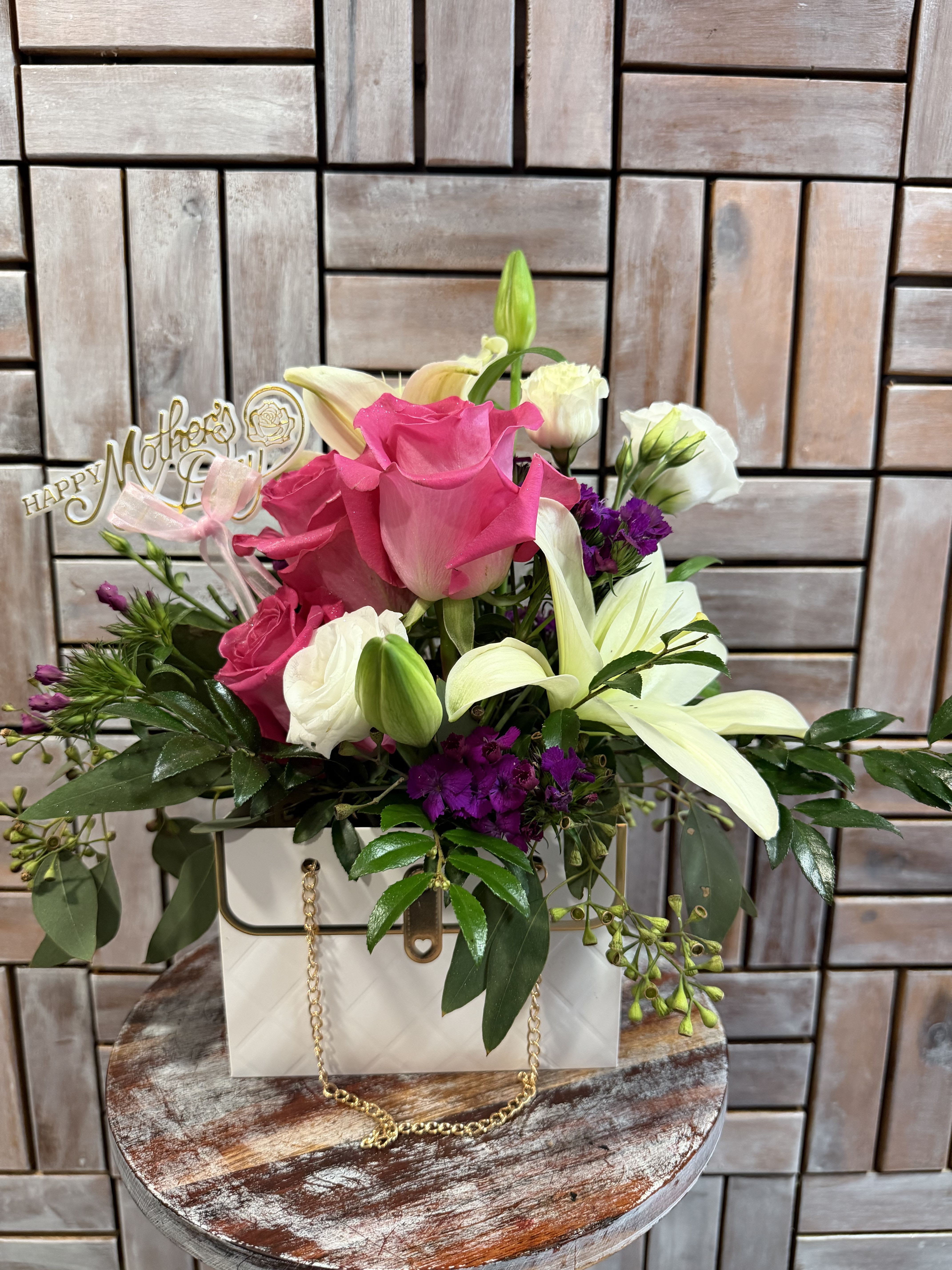 Elegance Bloom Box - A beautiful purse-inspired floral arrangement featuring vibrant pink roses, creamy white lilies, delicate lisianthus, and rich purple accents, all nestled in lush greenery. Designed in a stylish keepsake box with a gold chain detail. This arrangement is the perfect blend of elegance and charm—thoughtful, modern, and ready to brighten any day.