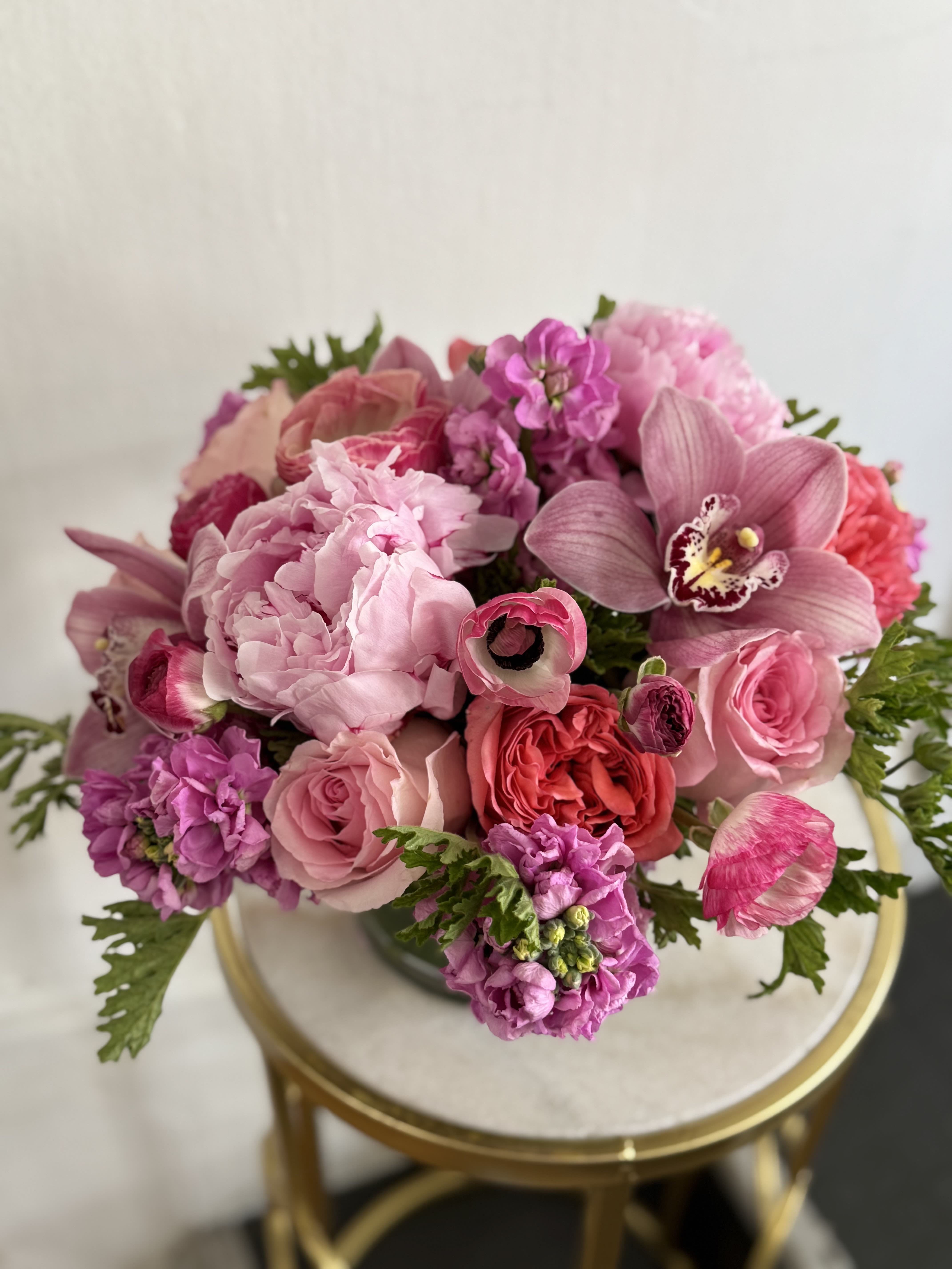 Premium Pink Please  - A lush, compact, tightly gathered arrangemen a palette of  layered pinks from whispery blush to hot magenta. It sets the tone for this romantic flower arrangement, perfect as a birthday bouquet, anniversary roses alternative, or a spring celebration gift. The recipient will feel deeply cherished as they take in the soft, full peony-like blooms and classic rose heads that create a sumptuous, feminine center, while sculptural orchid petals lift the design with elegant flair.  Choose this arrangement when you want someone to feel truly seen-whether you're saying "happy birthday," celebrating an anniversary, or just sending a thoughtful springtime surprise. The size and dense bloom count give the bouquet immediate visual impact; simple care-fresh water and a cool spot away from direct sun-will help the flowers last through the moment you've made for them.