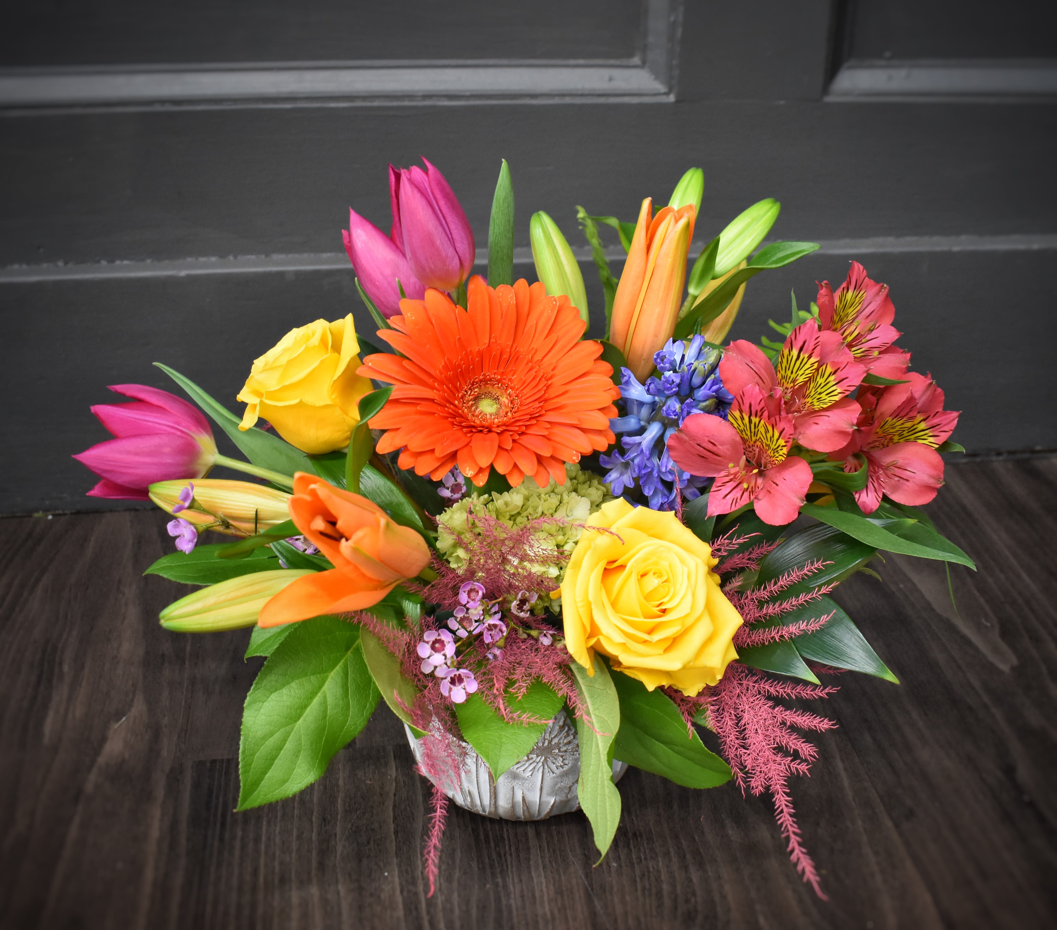 Burst of Happiness - PREMIUM - A vivid centerpiece that reads like a joyful exclamation - this mixed-color flower arrangement is a birthday bouquet, thank-you gift, or surprise "just because" pick-me-up all rolled into one. The person you're thinking of will feel instantly celebrated by the lively mix of orange, yellow, pink and purple tones and the bright, uplifting presence of a low silver vase that keeps the design fresh and table-ready.  The composition features a large orange gerbera daisy anchoring the center, surrounded by yellow roses, pink tulips, orange lilies, pink alstroemeria and blue‑purple hyacinth for surprising contrast and texture, with lush hydrangea massing for volume. Dense, layered and thoughtfully balanced, it offers a cheerful, tactile mix of petal shapes and scents that reads as both casual and premium.  Perfect for birthdays, thank-yous, or brightening someone's day, this arrangement sits low in a sleek silver vase so it works well on dining tables or desks without blocking conversation. 