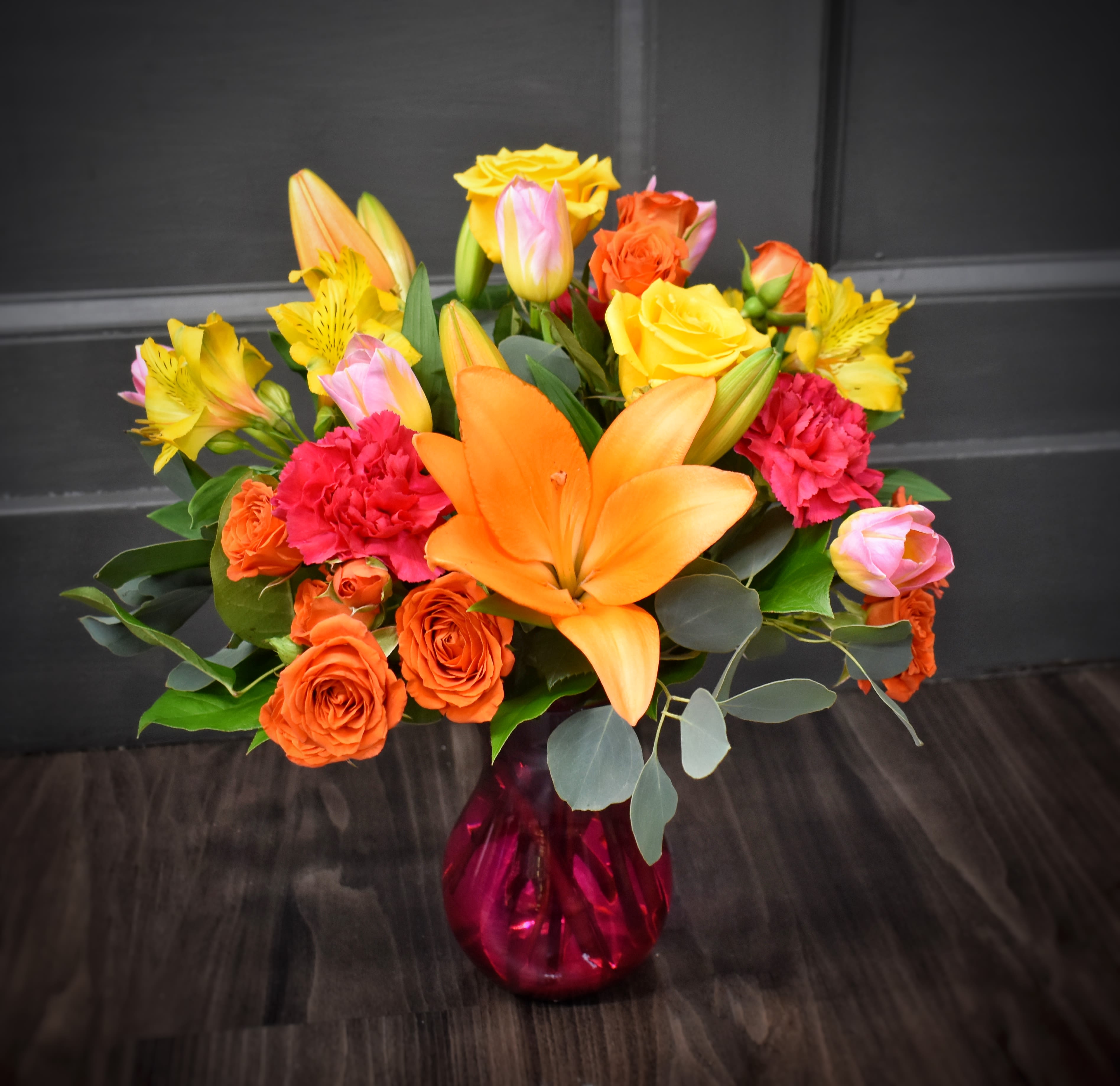 Sunny Disposition - PREMIUM - Sunlit orange spray roses and lilies take center stage in this cheerful flower arrangement, a bright birthday bouquet or thank-you gift that will make the person you're thinking of feel genuinely celebrated. Warm yellow roses and alstroemeria mingle with soft pink carnations and lush greenery for a layered, rounded composition that reads festive and uplifting-perfect for everyday gifting or to mark a happy occasion.  The blooms are artfully gathered at varied heights for a full, lively silhouette and arrive ready in a bold red glass vase that gives the bouquet a gift-ready pop. Colors: orange, yellow, pink and green. Flowers: roses, lilies, carnations and alstroemeria. The glass vase both anchors the arrangement and reflects the sunny palette-no extra wrapping needed. For best enjoyment, keep in fresh water, trim stems if needed, and place out of direct heat to extend the cheerful display.