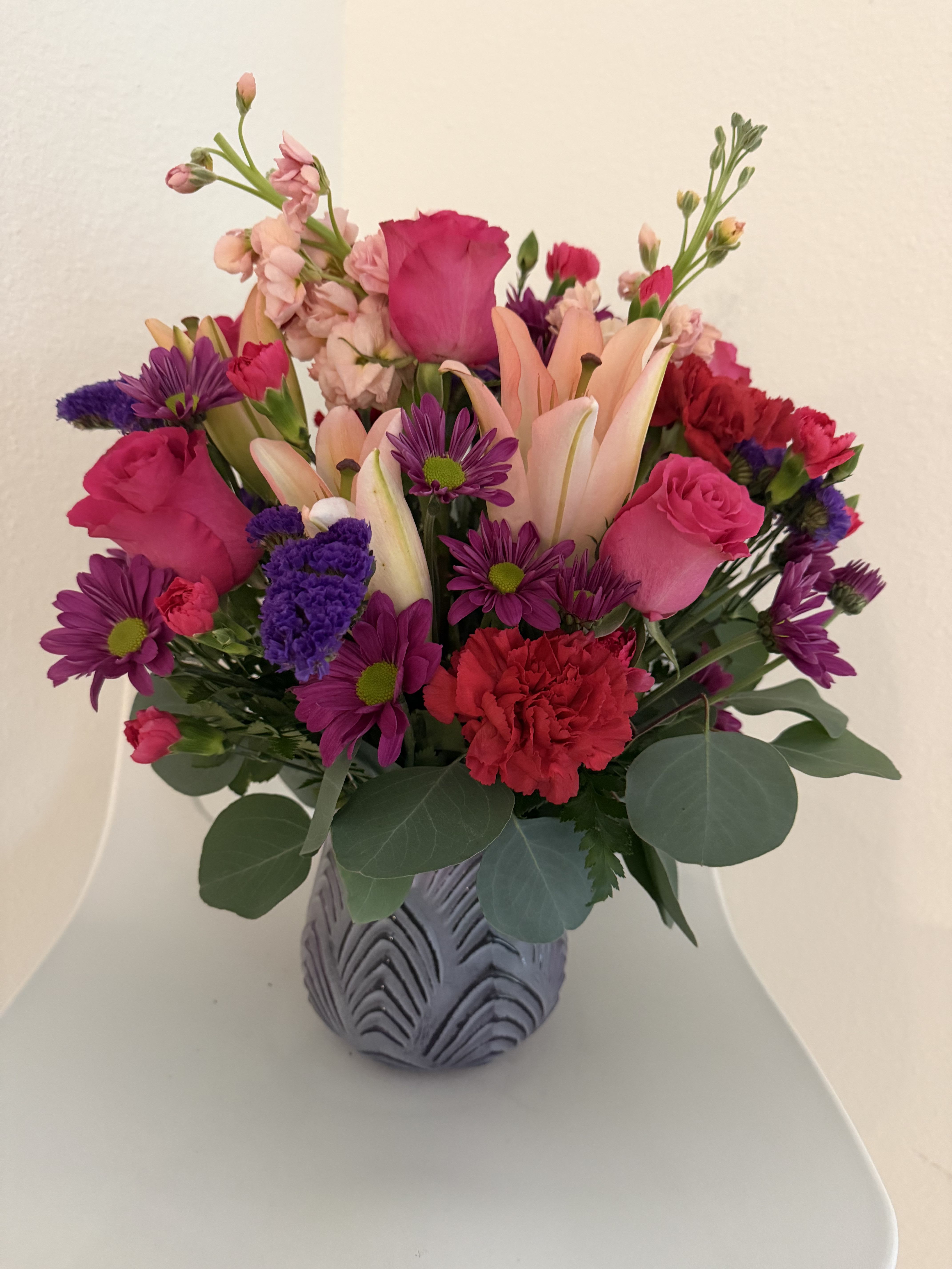 Blushing Garden Harmony - A vibrant and elegant arrangement featuring pink roses, soft peach lilies, purple daisies, and red carnations, beautifully complemented by fresh greenery in a modern textured vase. Perfect for brightening any space with a touch of warmth and charm.