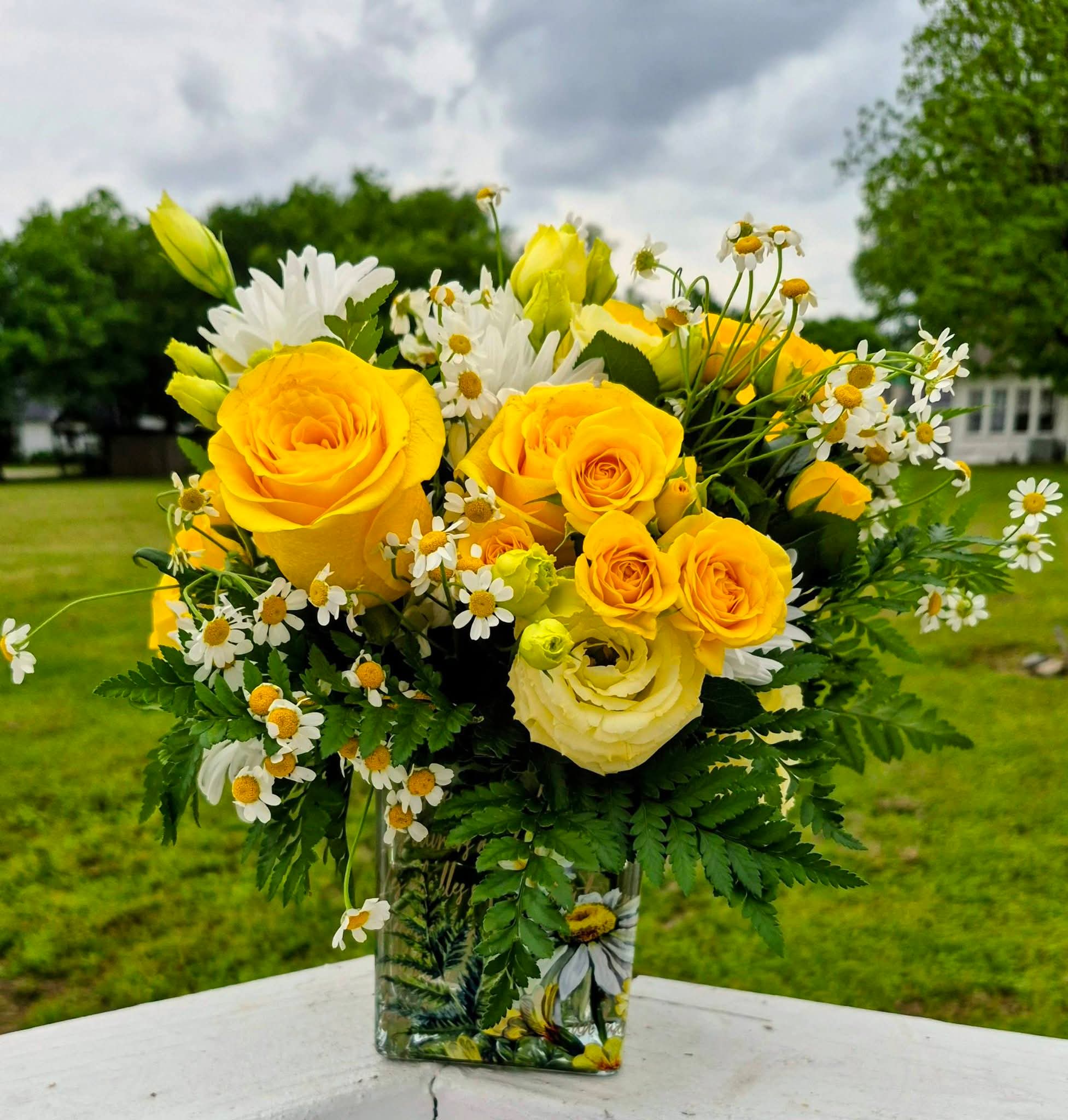 BEE-UTIFUL - Bring a breath of fresh air indoors with this cheerful, garden-inspired arrangement bursting with warm yellow tones and crisp whites. Featuring radiant yellow roses, delicate spray roses, and soft lisianthus, this design is perfectly balanced with classic white daisies and airy chamomile for a light, whimsical touch. Lush greenery adds texture and movement, creating a natural, just-picked-from-the-garden feel.