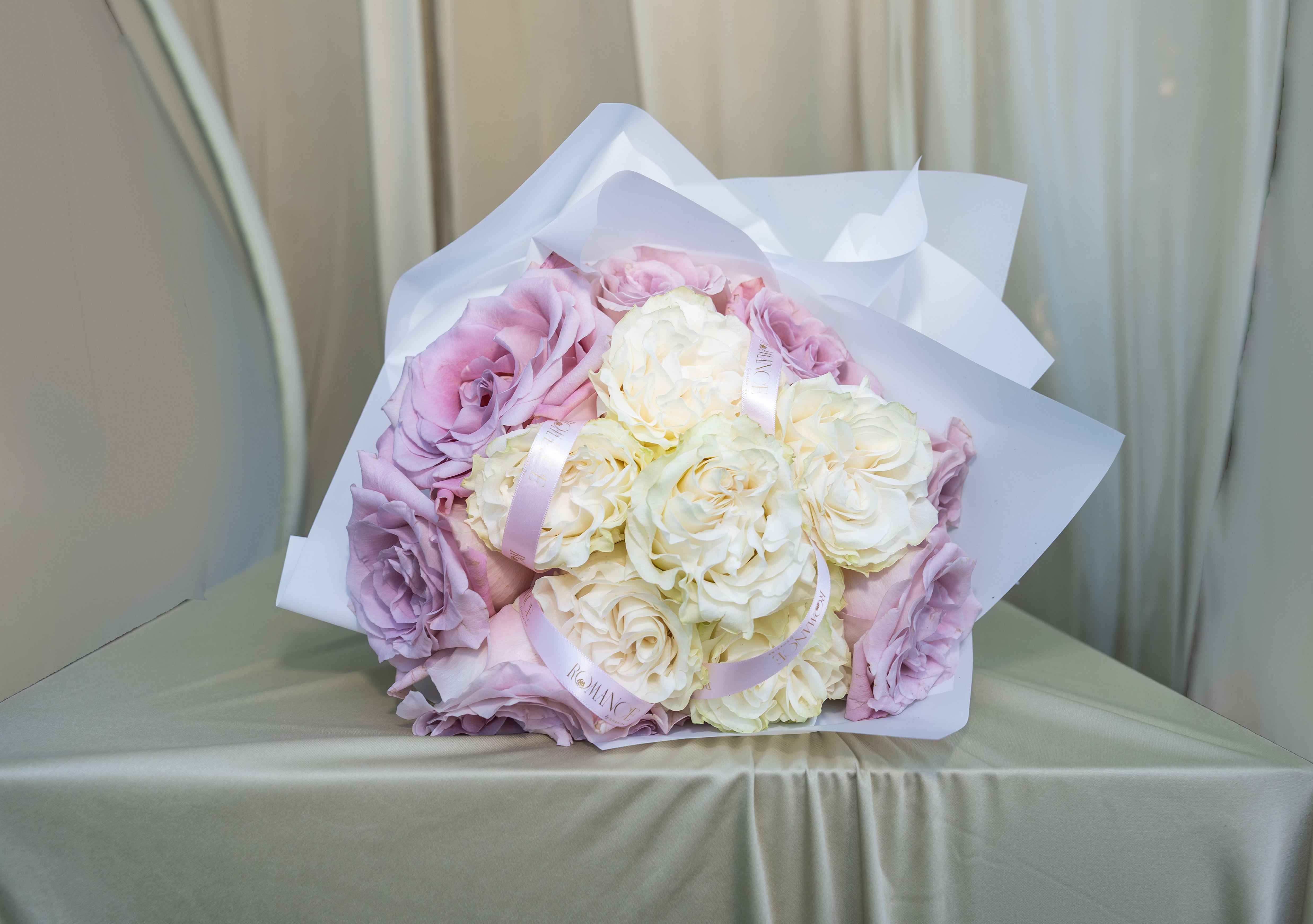 The Beloved - Some women don't need to be told they are loved. They feel it in the details… in the softness of what is chosen for them, in the care woven into every layer. The Beloved was made for her. A lush, generous gathering of dusty mauve roses and creamy ivory garden roses, wrapped in soft white paper and finished with a Romance by Shakira ribbon woven intimately through each bloom. Nothing is accidental here. The ribbon doesn't just adorn the bouquet… it moves through it, the way love moves through a life, quiet and constant and present in every layer. The mauve is moody and tender. The ivory is soft and luminous. Together they speak the language of devotion without ever needing to raise their voice. This is the bouquet you give to the woman who means everything… the one who nurtured a family with her whole heart, who loved deeply and endlessly, who has always been, in every sense of the word… beloved. Whether she is your mother, your partner, or the woman who made you a family… she deserves to feel it today. Give her something that knows her name.