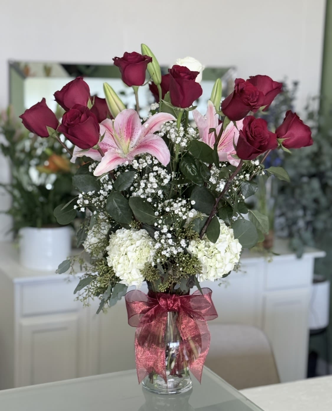 1Dz Red Roses With White Hydrangeas And Pink Asiatic Lilles. - Tall red roses rise above soft pink lilies in a romantic birthday bouquet built for anniversaries, Valentine's Day, or any moment when you want your person to feel truly cherished. The upright composition pairs deep red and blush pink for a classic love-and-celebration contrast, while white hydrangea and delicate white fillers soften the silhouette so the arrangement reads both lush and intimate.  Fresh roses, fragrant lilies, and full white hydrangea are artfully arranged with greenery for texture and height. Presented in a clear glass vase and finished with a sheer burgundy ribbon, the arrangement has a polished, gift-ready look that suits a special dinner, a bedside surprise, or a thank-you that feels memorable. Size and vase dimensions are visible on the product page; water daily and trim stems every few days to keep blooms looking their best.