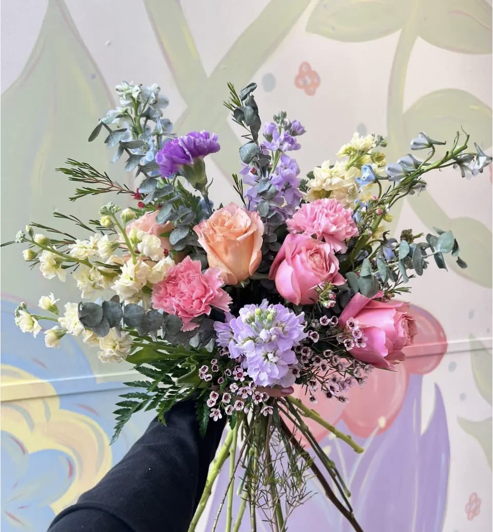 Burst of Blooms - Hand Tied - Soft pastel romance in a loose, garden-style hand-tied bouquet-perfect as a birthday bouquet, anniversary flowers, or a thoughtful everyday gift for someone who loves gentle color and natural charm.   This arrangement features long, visible stems tied into an airy, informal composition with blue-gray eucalyptus and fine-textured greenery for contrast and texture. The palette of pink, purple, peach, and white offers layered softness and a light, fragrant presence from the roses and stock. Finished in a casual hand-tied style, it reads as newly gathered from the garden-bright without being fussy.  Its relaxed, natural presentation makes it a meaningful and effortless way to lift someone's day, especially for someone who has a special vase at home.