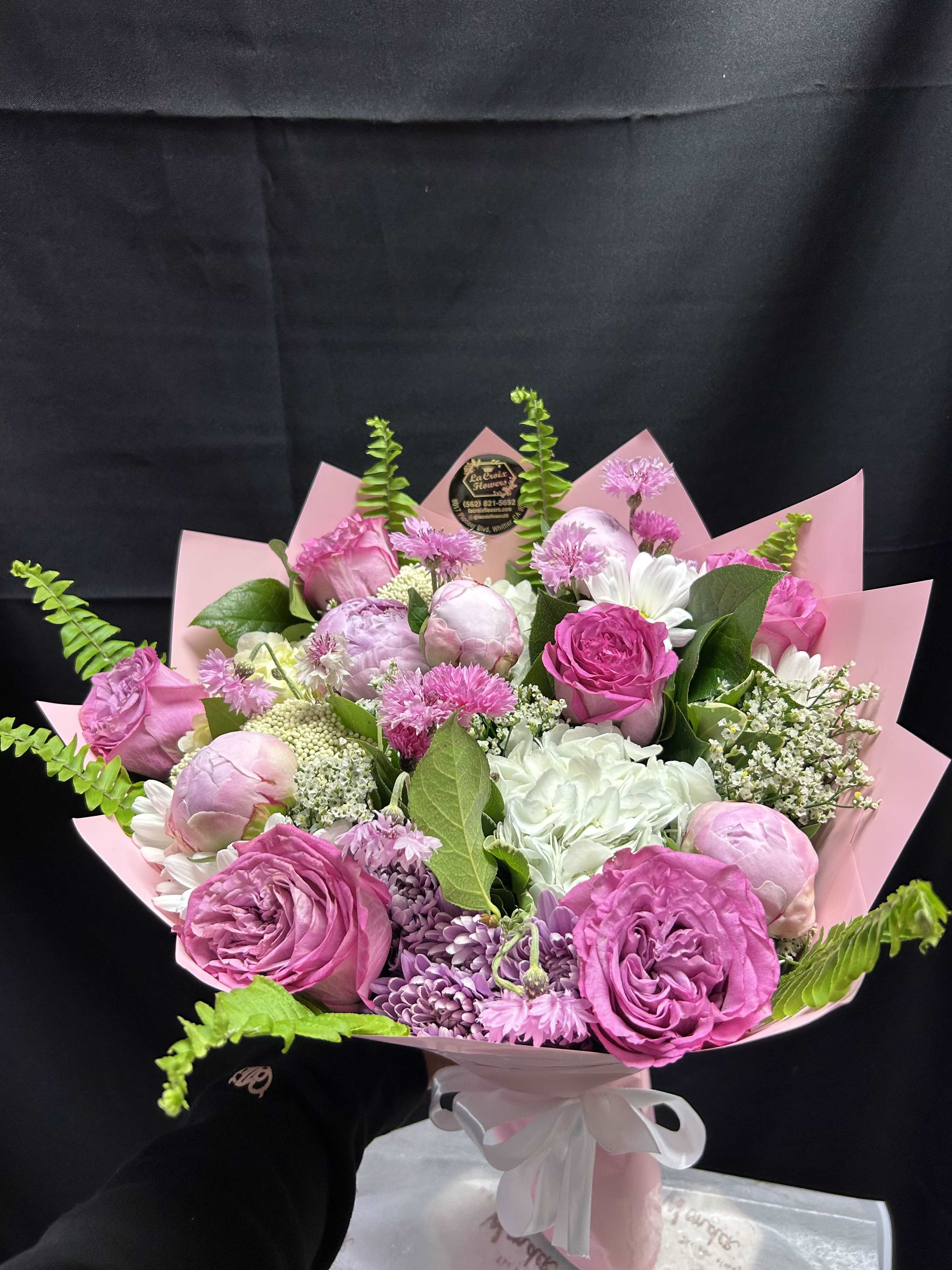 Love Pionies (mixed bouquet) - Soft blush peonies and bright pink roses steal the show in this hand-tied bouquet-perfect as a birthday bouquet, anniversary roses gift, or a Mother's Day surprise. The lush, rounded arrangement reads romantic and garden-fresh at first glance, with purple chrysanthemum-like blooms and white daisies adding contrast and a touch of playful texture that makes the recipient feel genuinely celebrated.  Packed with peonies, roses, daisies, chrysanthemums, carnations, and clustered white filler flowers, the palette blends pink, white, purple, and fresh green for a full, feminine look. Finished in smooth pink wrapping paper and tied with a white satin ribbon plus a floral label, the presentation is gift-ready and polished. Bright green fern fronds weave through the stems to add movement and a lively pop of color.  Ideal for marking birthdays, anniversaries, spring celebrations, or simply saying "I'm thinking of you," this bouquet arrives ready to impress. Treat the person you care about to a soft, fragrant arrangement that looks as though it were just picked from the garden; ordering notes and sizing are shown on the product page for easy customization.