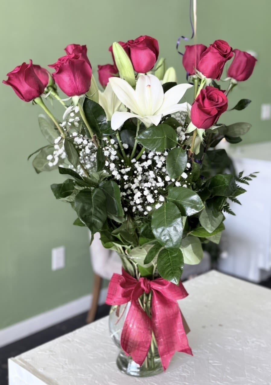 1 Dz Re Roses In A Vase - a dz of  red roses circling a single white lily creates a striking focal point-this upright flower arrangement is a perfect anniversary bouquet or birthday surprise that reads as both romantic and celebratory. The vivid pink-and-white contrast and glossy green foliage make it an ideal gift for love and romance, Valentine's Day, Mother's Day, or a "just because" moment when you want her to feel truly cherished.  Fresh redroses and a central white lily are accented with delicate white baby's breath and layered greenery for a full, textured look. The stems sit in a clear glass vase, finished with a bright pink ribbon and decorative wrap for a polished presentation. Sized to sit beautifully on a dining table or bedside dresser, this arrangement arrives ready to display-trim stems, change water every two days, and remove any wilted foliage to keep blooms looking their best.