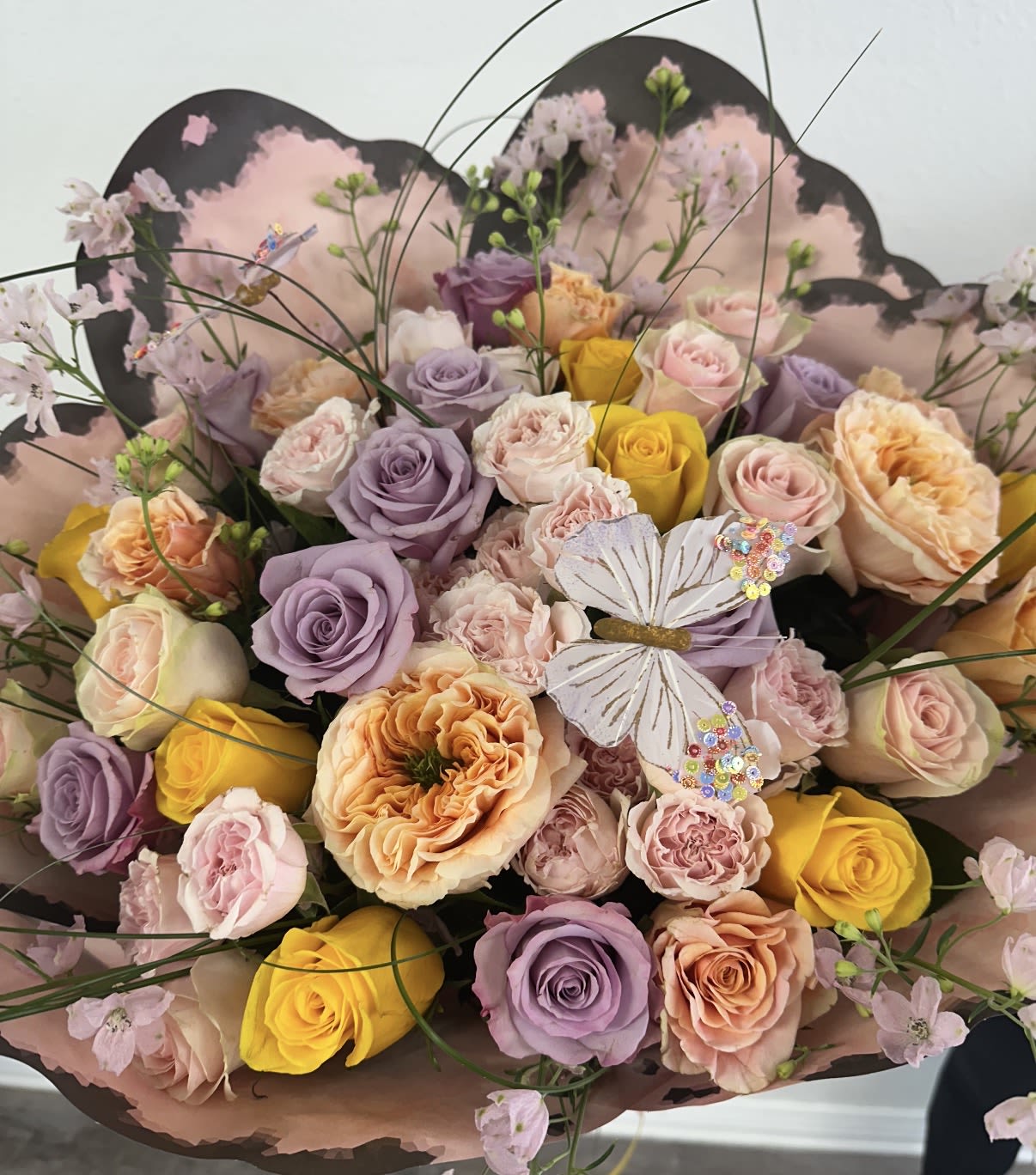 The Butterfly Meadow - Capture the essence of an enchanted garden with this lush, garden-style bouquet. Featuring a harmonious palette of lavender, apricot, and sunny yellow roses, this arrangement is textured with delicate spray roses and airy delphinium accents.  The centerpieces of this design are the exquisite David Austin-style garden roses, known for their high petal count and ruffled, romantic appearance. Nestled among the blooms is a handcrafted sequined butterfly embellishment, adding a playful touch of magic.