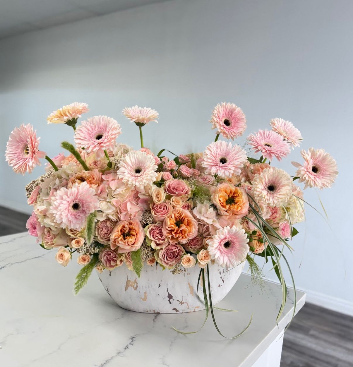 The Peach Prosecco - Elevate any space with this luxurious, low-profile centerpiece that radiates warmth and modern charm. Presented in a distressed white and gold artisan bowl, this arrangement features a rich foundation of blush hydrangeas and velvety spray roses in varying shades of peach and apricot.  The true stars of this design are the dancing gerbera daisies, which are artfully "floated" at different heights to create a sense of movement and whimsy. Accented with trailing greenery and delicate filler stems, this piece offers a 360-degree view of floral elegance, making it an ideal choice for dining tables, kitchen islands, or executive desks.