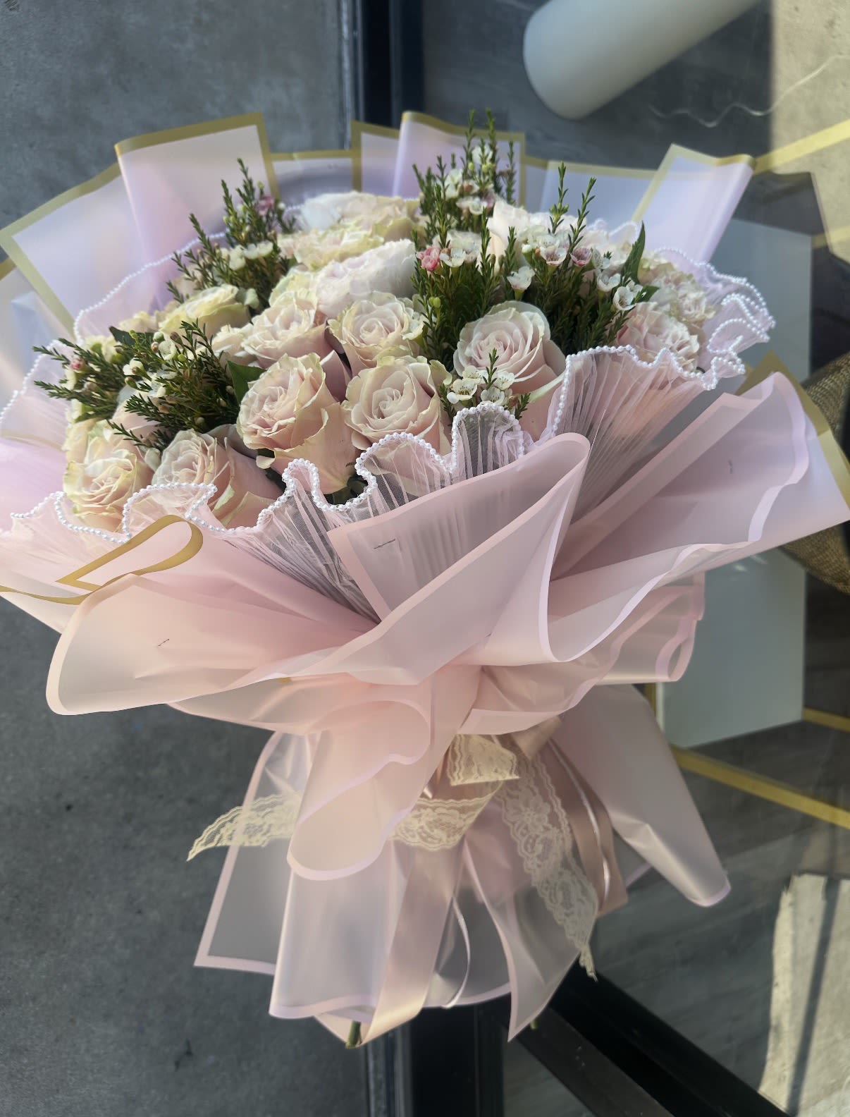 The Pink Pearl Reverie - Indulge in pure romance with our Pink Pearl Reverie, a bouquet designed for those who love the finer details. This arrangement features a stunning collection of creamy blush roses, chosen for their soft, spiral petals and timeless elegance. Tucked between the blooms are sprigs of fresh waxflower, adding a delicate, starry texture and a light, citrusy fragrance.  What truly sets this bouquet apart is its exquisite "couture" presentation. The roses are nestled in layers of soft blush waterproof wrap, accented by a unique pleated white tulle border tipped with miniature pearls. Finished with a vintage-style lace ribbon, this bouquet feels like a hand-delivered love letter.