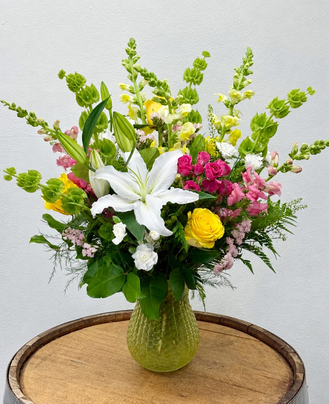 Love, Mom's Favorite - A dramatic white lily rises at the heart of this cheerful flower arrangement, surrounded by sunny yellow and hot-pink roses-perfect as a Mother's Day bouquet or a "just because" arrangement to settle (or provoke) a bit of sibling rivalry with style. The bright mix of green, white, pink and yellow reads celebratory and welcoming, so the person you're thinking of will feel genuinely noticed and loved.  Pink snapdragons and ruffled carnations add vertical movement and texture while delicate white and blush filler blooms soften the palette; tall green bells of ireland lift the composition into a garden-like, upright silhouette. Everything sits in a textured yellow glass vase that gives the design a sunny day charm and keeps it sized nicely for tabletops and entryways.  This is the kind of arrangement that says "she's special"-for Mother's Day, birthdays, thank-yous, or any spring moment when you want to make Mom feel celebrated. It brightens a room and holds its shape with regular water changes; order early to ensure it arrives fresh for the big day.