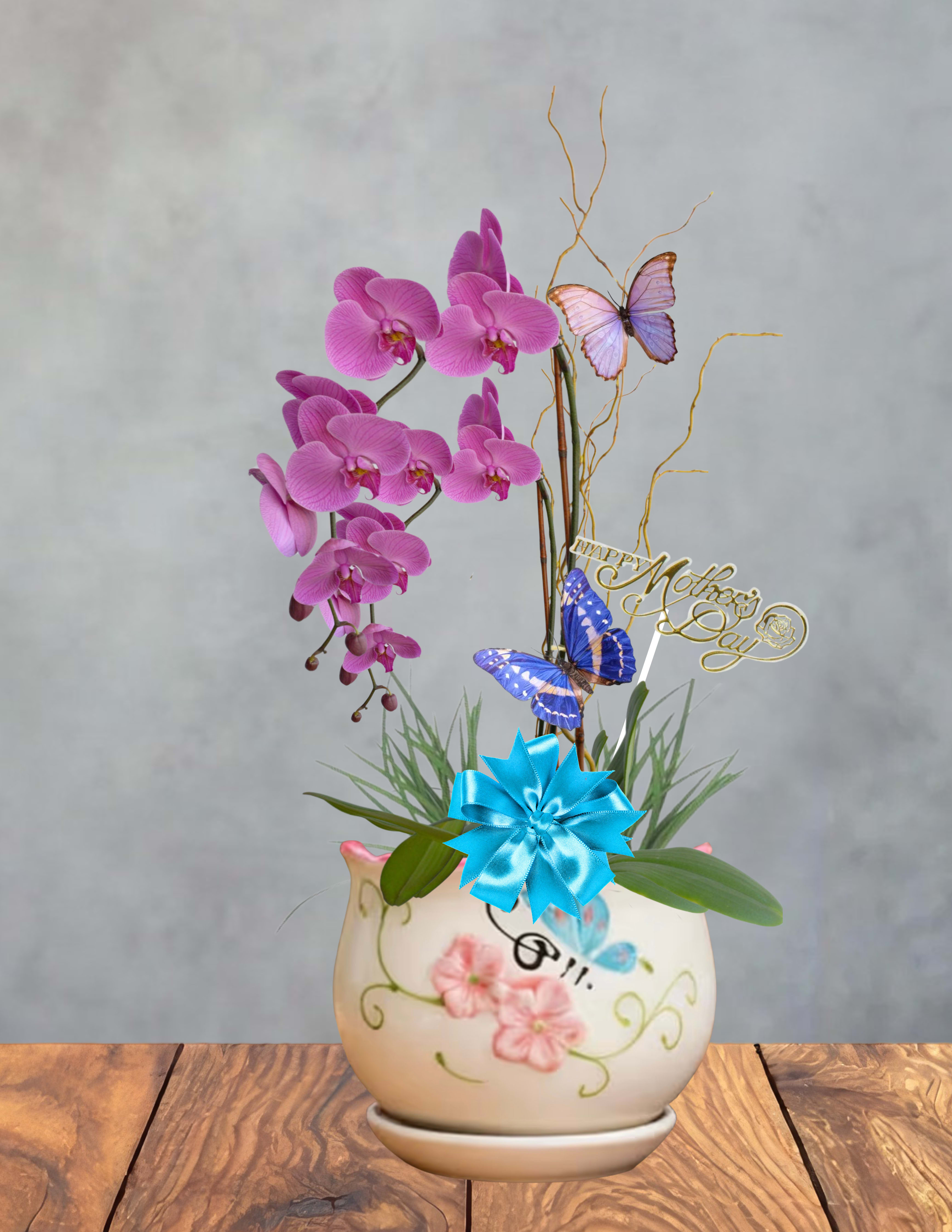 Orchid butterfly garden (color may vary)  - Soft pink Phalaenopsis orchids arch gracefully above a painted rounded ceramic vase, creating a delicate Orchid butterfly garden perfect as a Mother's Day flower arrangement or a cheerful birthday gift for someone who loves elegant blooms. The feminine palette of pinks with possible accents of purple, blue and white and the fluttering decorative butterflies give the recipient a feeling of being celebrated and adored-this is the kind of gift that makes her feel truly seen.  Several stems of pink Phalaenopsis rise from the vase with thin curly branches that add height and an airy texture, finished with a bright blue ribbon bow and butterfly picks for a whimsical touch. A metal pick topper (shown with a gold "Happy Mother's Day" message) and the hand-painted floral ceramic container give the display a polished, gift-ready presentation.  Ideal for moms, grandmothers, teachers, or any special woman who appreciates lasting orchids and pretty keepsakes; they'll enjoy the long-lasting blooms and the decorative vase that can be reused. Colors may vary slightly by availability; care for Phalaenopsis orchids by placing in bright, indirect light and watering sparingly to keep blooms looking their best.